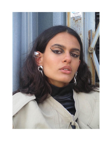 Woman with short, dark hair leans against wall, wearing silver chain earrings