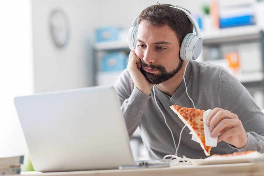 Lazy man sitting at desk using a laptop and having a ready meal he is holding a slice of pizza