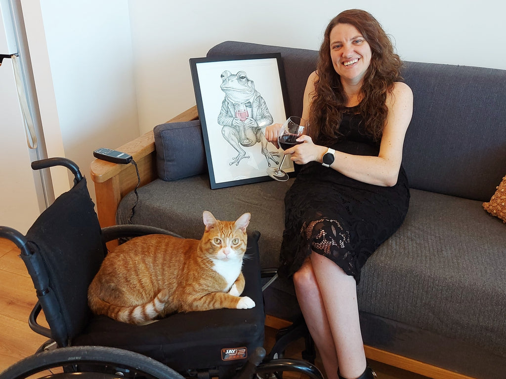 Cobie sitting on a lounge drinking a glass of wine next to the Jeremiah drawings. Charlie the cat is sitting on Cobie's wheelchair in the foreground
