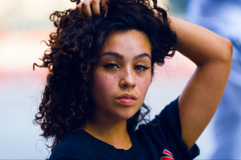 A woman with freckles and glowing skin holds her curly hair away from her face.