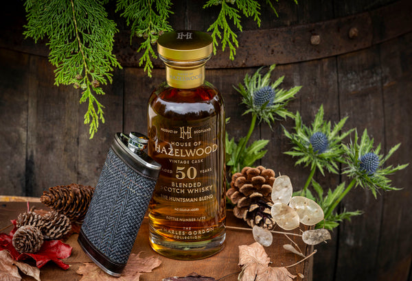 A bottle of The Huntsman Blend and Hipflask sit in front of a cask surrounded by thistles and pinecones.