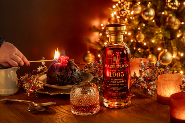 A picture of a bottle of Blended At Birth Whisky on a table set for Christmas. A hand holds a match ready to light the Christmas pudding.