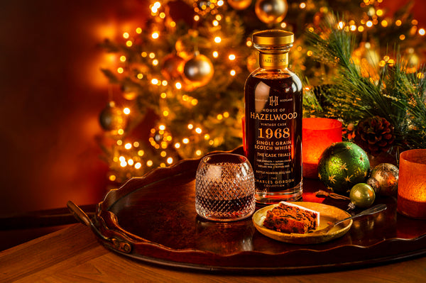 A bottle of The Cask Trials whisky sits on a walnut tray with a glass and a slice of Christmas cake. Baubles litter the table and a Christmas tree lights up the background.