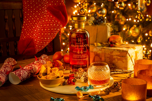 A bottle of The Old Confectioner's Whisky is surrounded by a Christmas stocking, humbug sweets and toffees with a Christmas tree in the background.