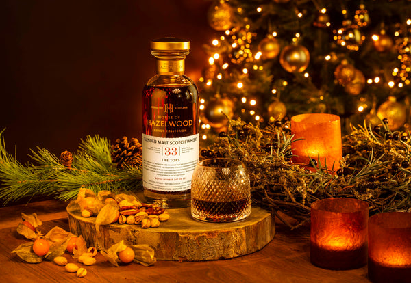 A bottle of The Tops Blended Malt Whisky sits atop a log slice, surrounded by seasonal nuts and fruit. A candle and a Christmas tree are seen in the background.