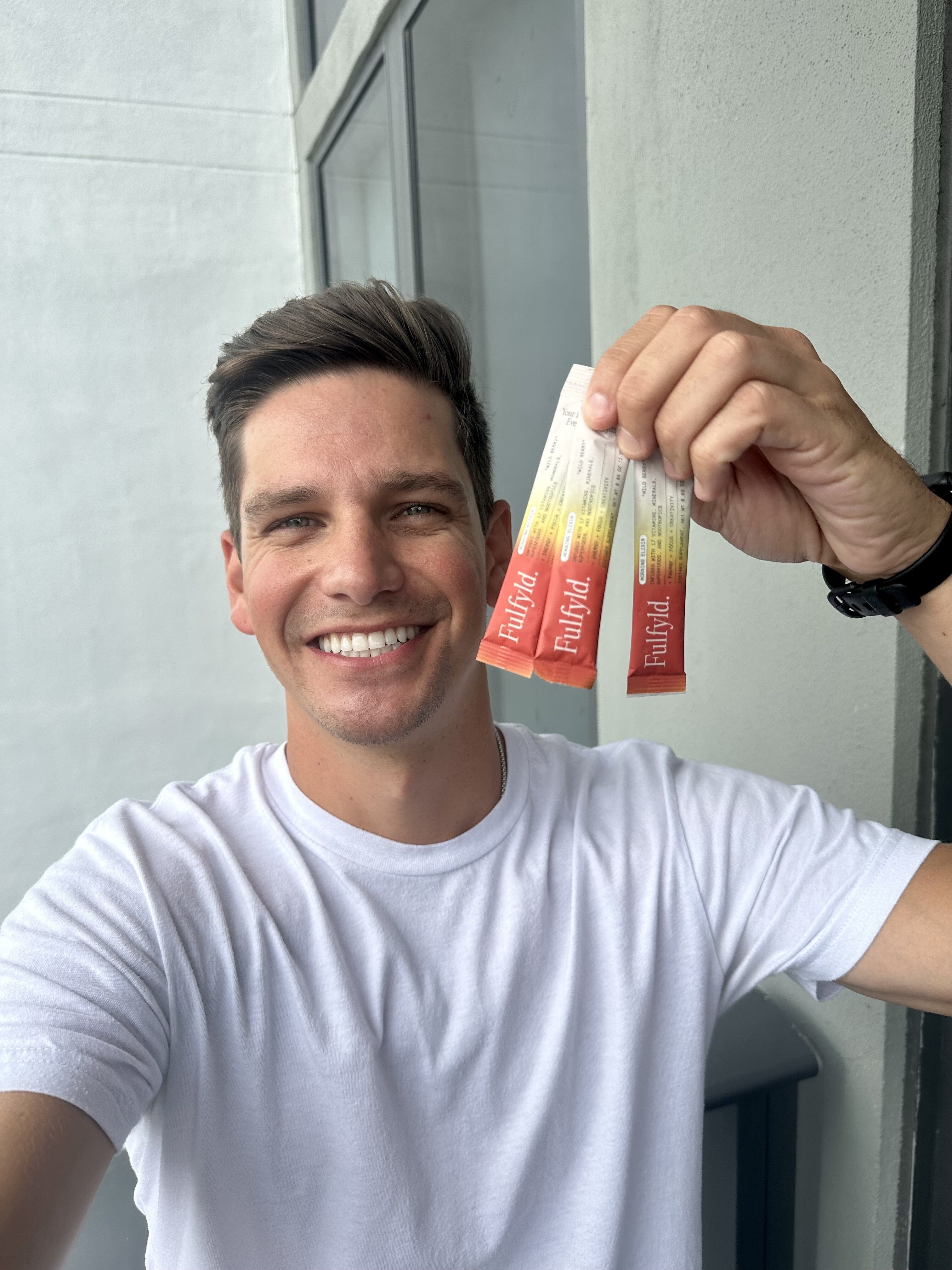 A person in a white shirt smiles while holding two FlavorAid packets.