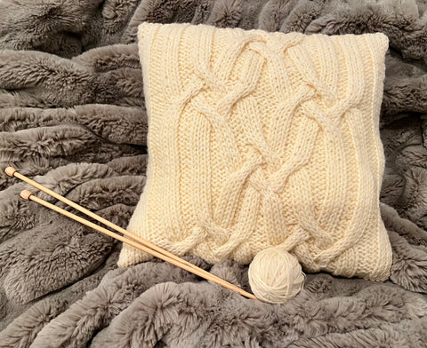 Friendship Cushion knit in white on a blanket, next to ball of yarn and needles.