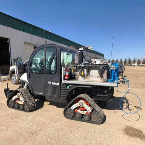 A black Bobcat utility vehicle on tracks, equipped with various mechanical tools and a blue hose, parked outside a green industrial building.