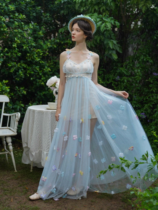 A woman in a sheer blue fairycore dress with pastel flower appliqués and pearl accents poses gracefully in a garden next to a tea table.
