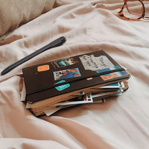 A much-loved Moleskine notebook sits closed on a bed, covered in stickers and bristling with photos stuffed inside its pages