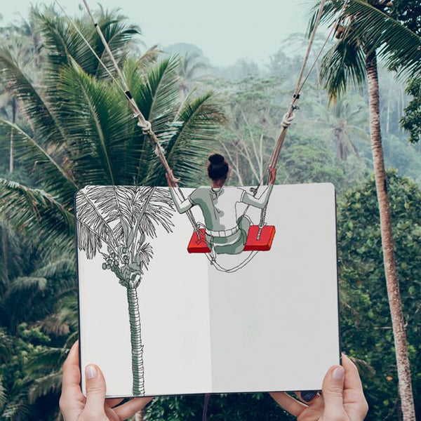 A drawn image in a notebook, of a tree and the lower half of a person on a swing, is held up so that it lines up with real trees and the person swinging
