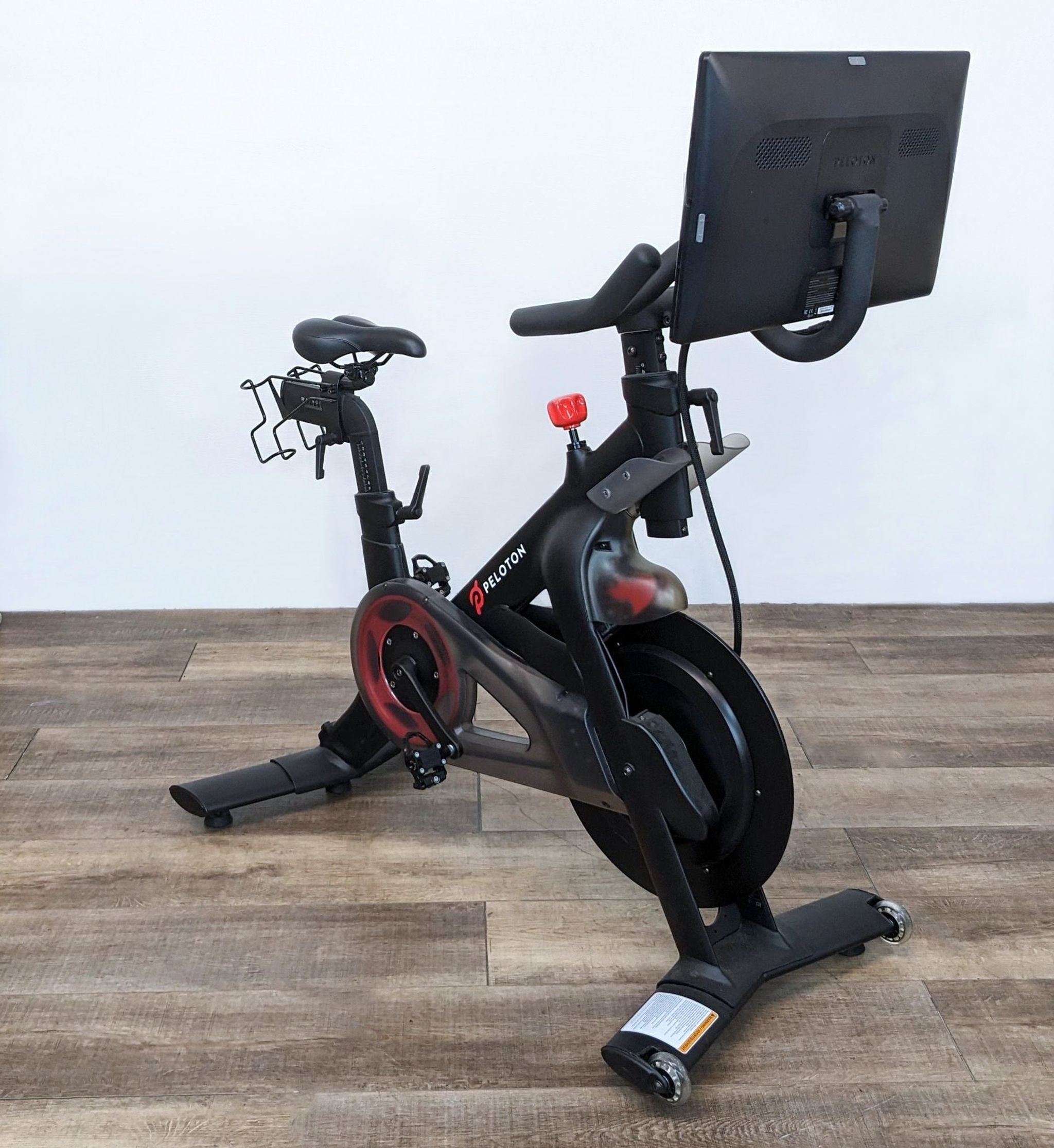 Side angle of a Peloton indoor cycling bike featuring branding, adjustable seat, handlebars, and a digital display monitor.