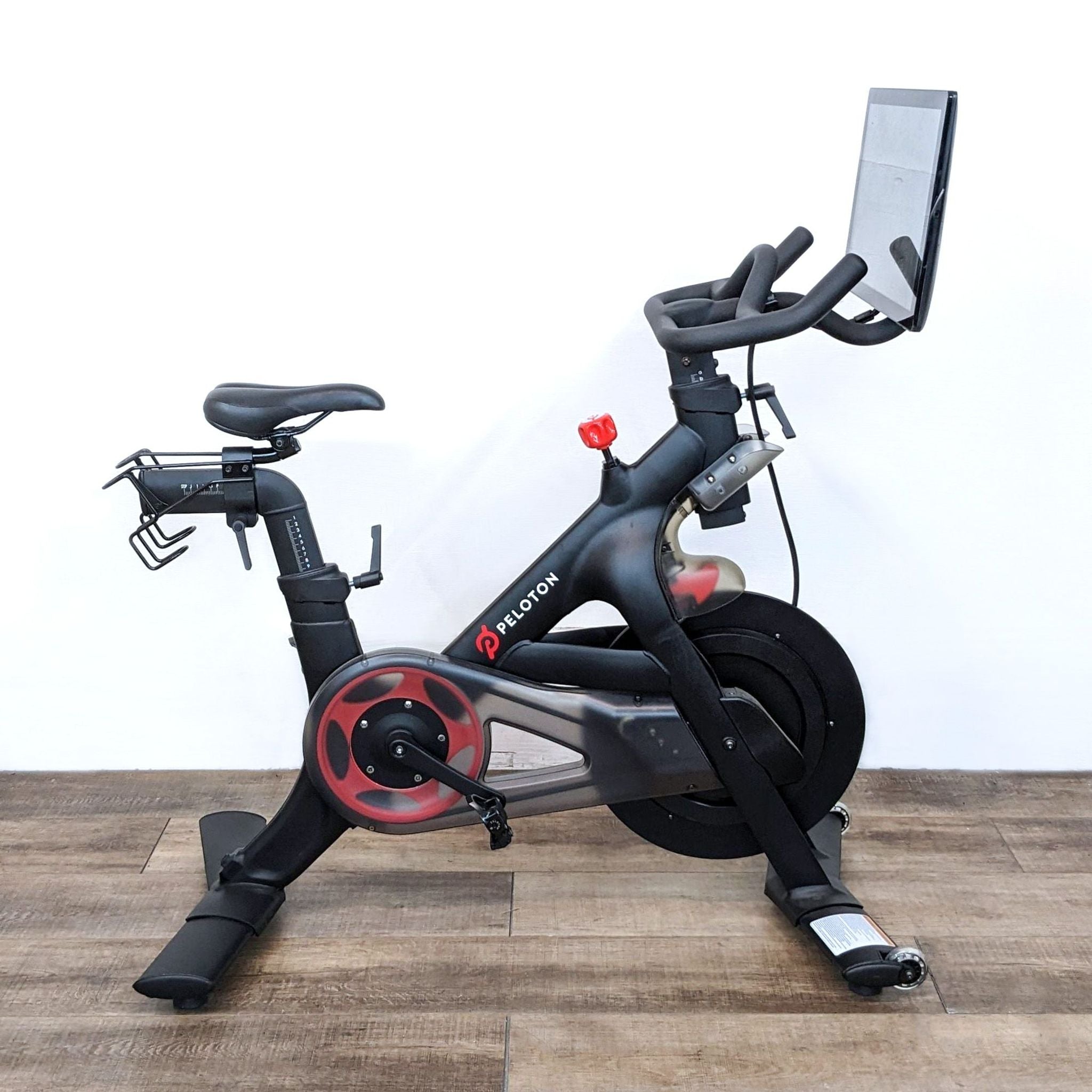 A Peloton exercise bike with a screen, set on a wooden floor against a white wall.