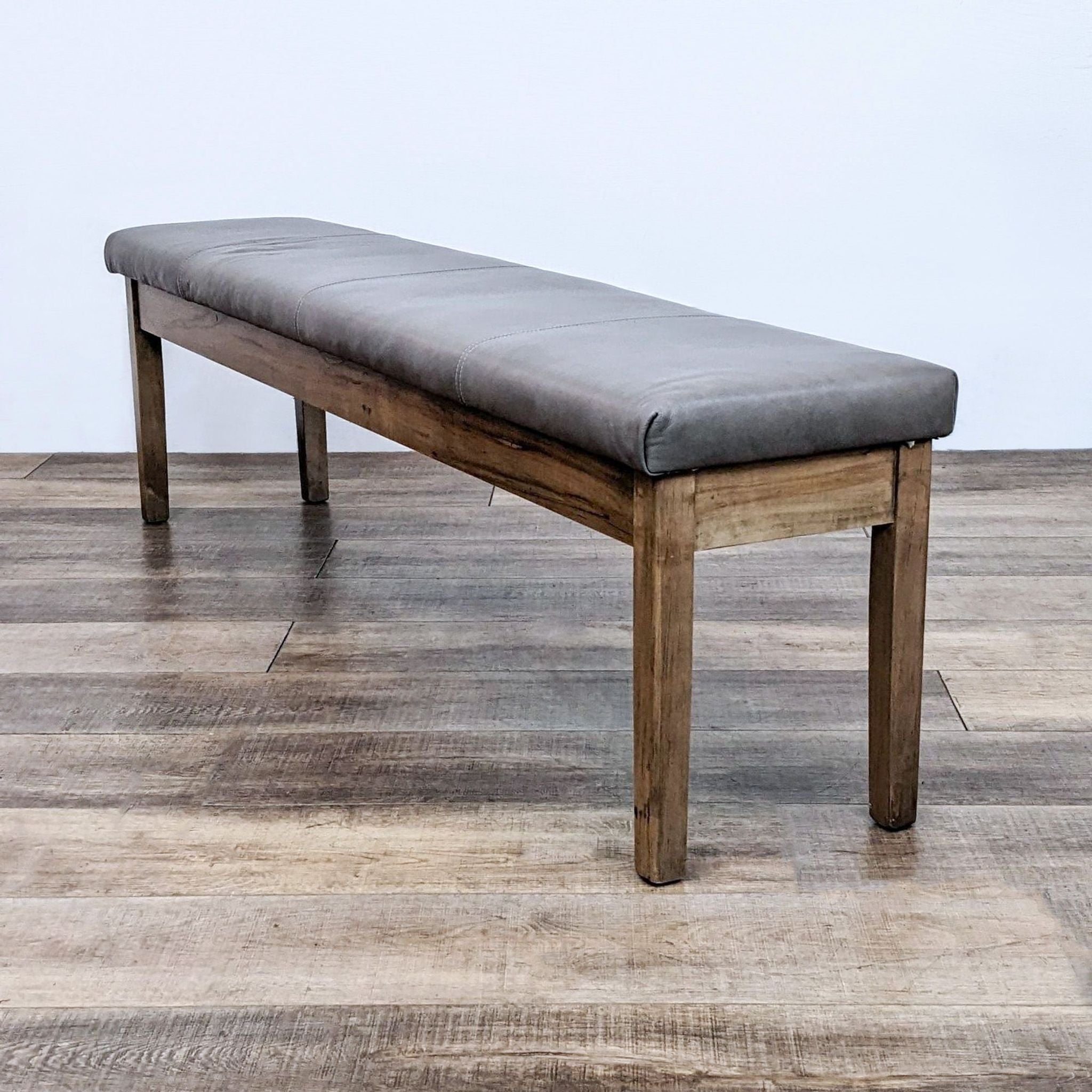 Alt text 2: A long, solid wood bench with a cushioned faux-leather seat by Bassett, positioned against a wooden floor backdrop.