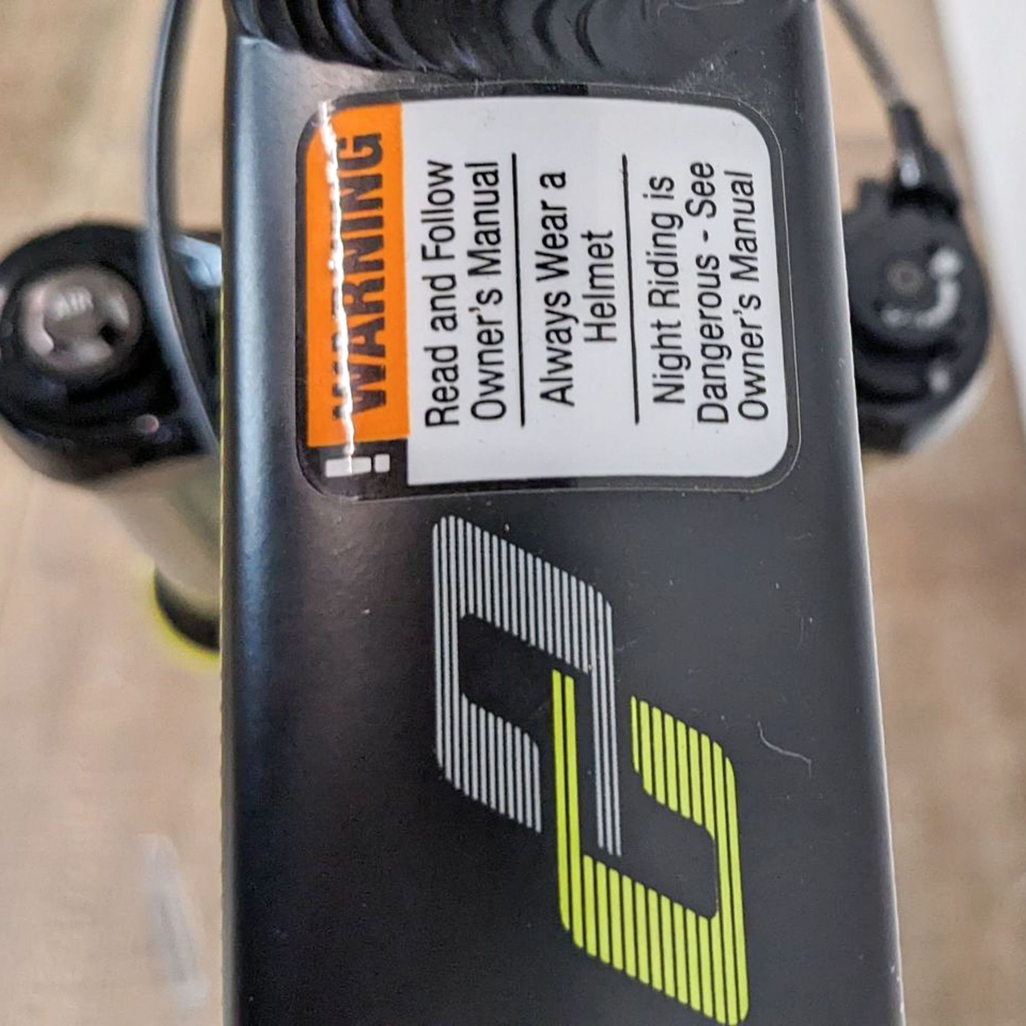 Close-up of a warning sticker on a bicycle frame with a black and yellow REI brand logo, advising to read the manual and always wear a helmet.