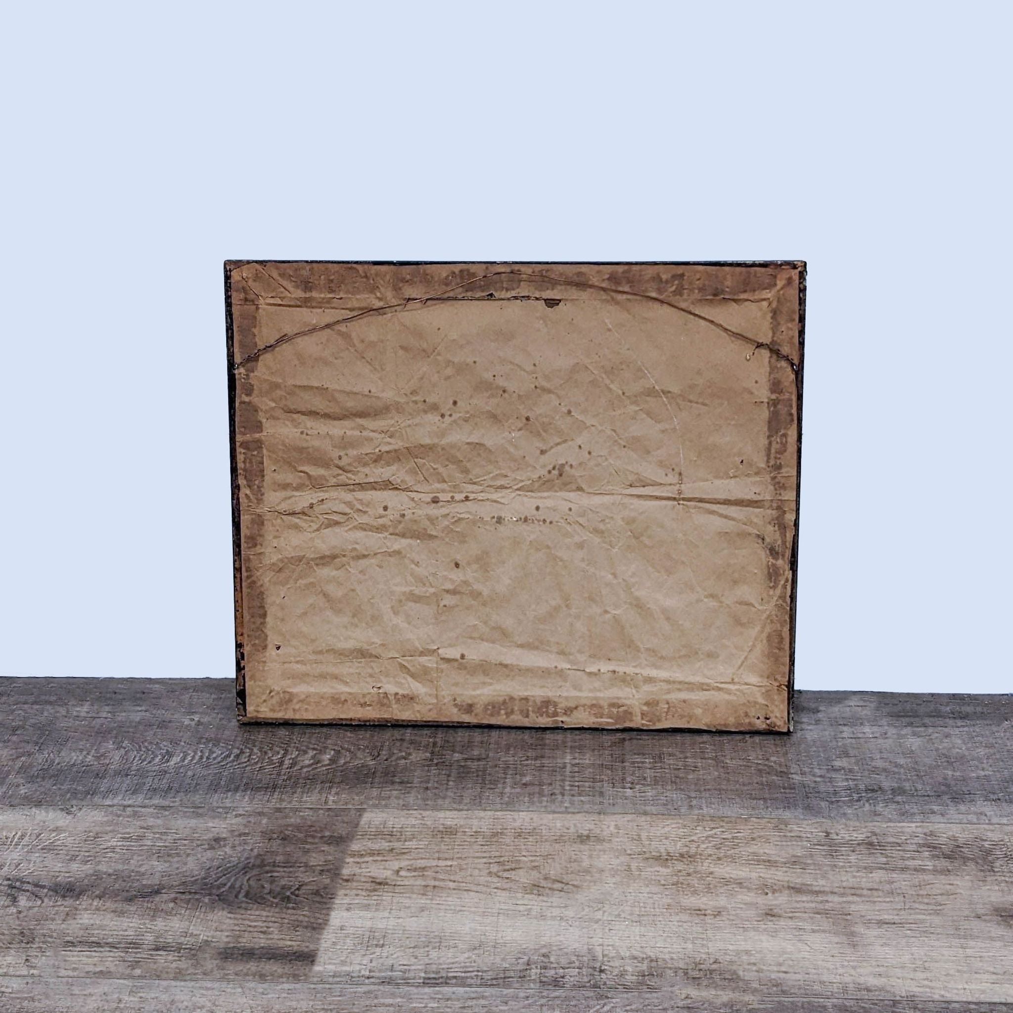 Back of a framed canvas showing aged paper and wire hanging assembly on a wooden surface against a white background.