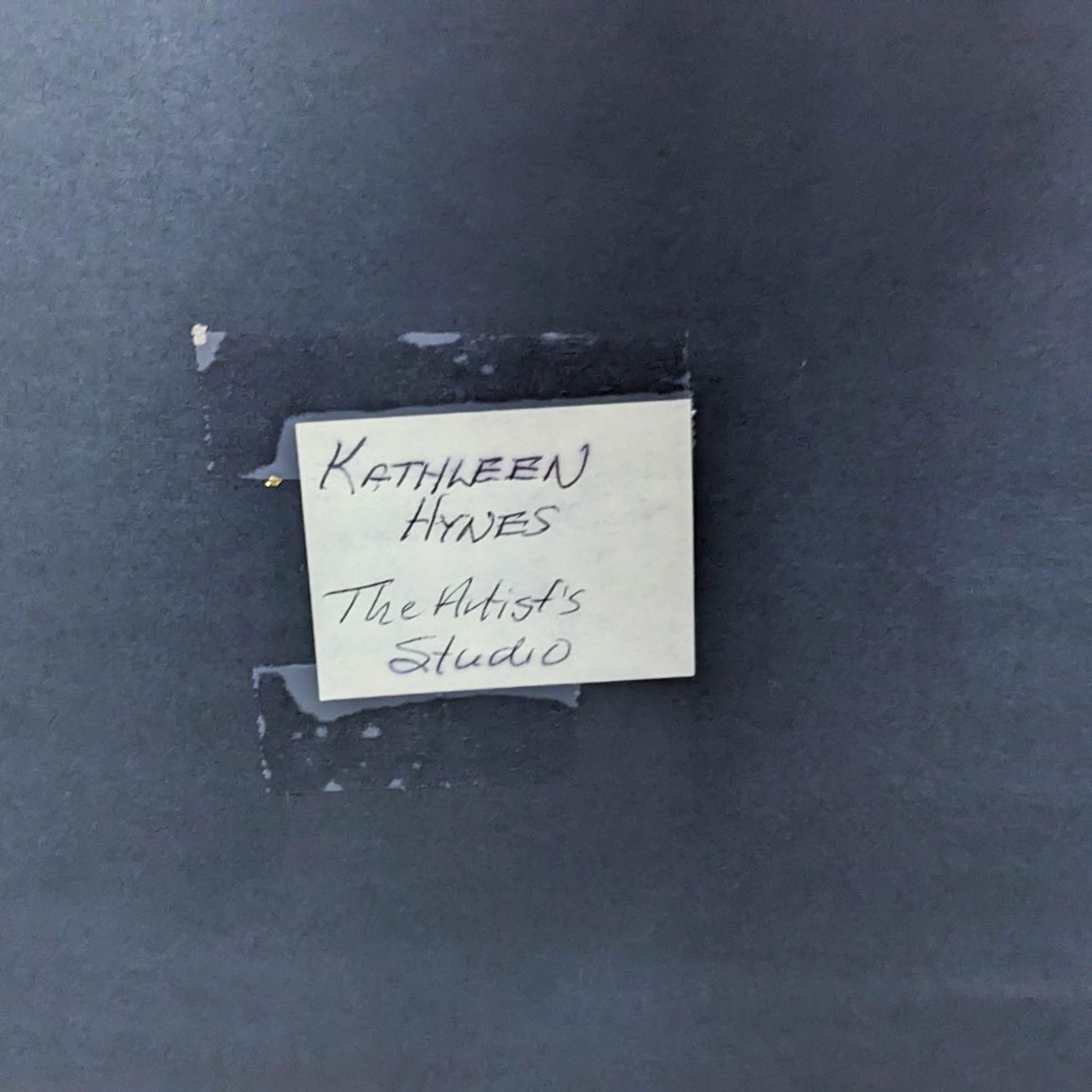 Piece of paper with "Kathleen Hynes The Artist's Studio" handwritten on it, taped on a dark surface.