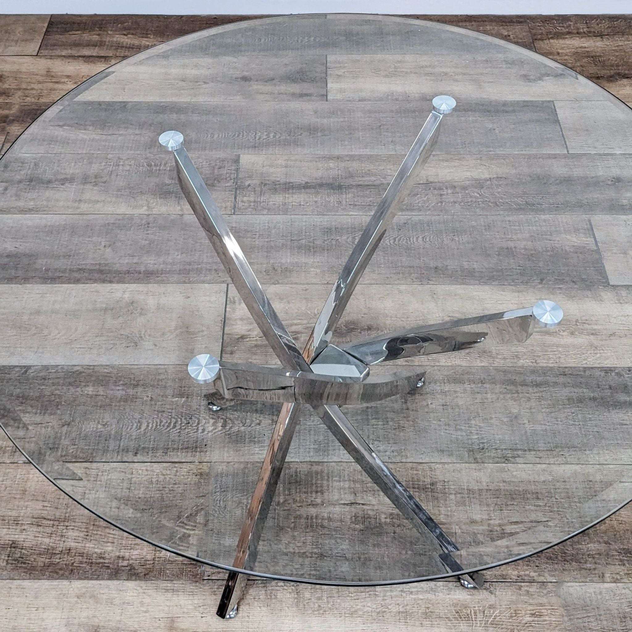 Top view of Reperch dining table showing the round glass surface and intersecting metal legs, placed on a wood-patterned floor.