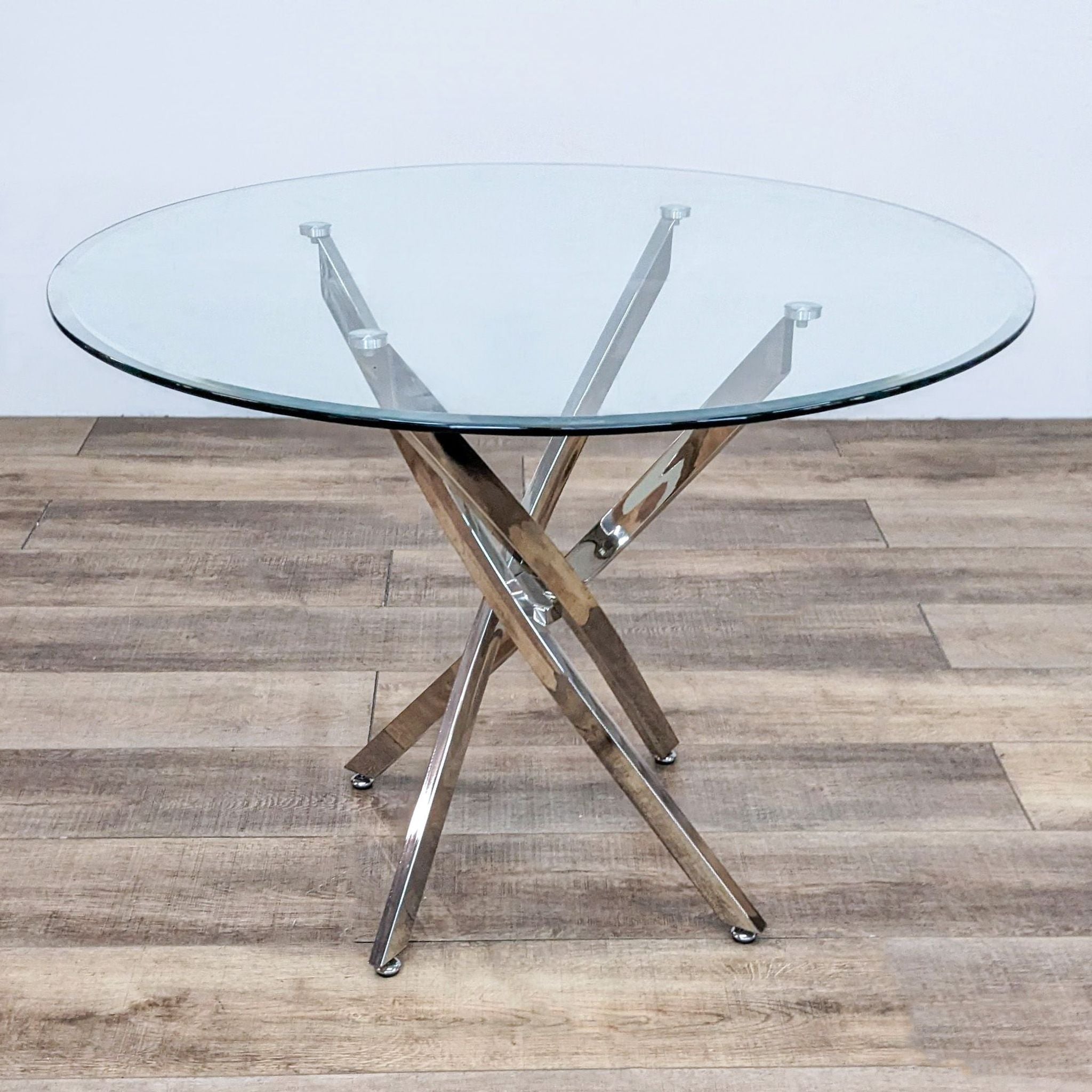 Reperch brand modern dining table with a thick clear tempered glass top and gleaming metal base with angled, slanted posts on a wooden floor.