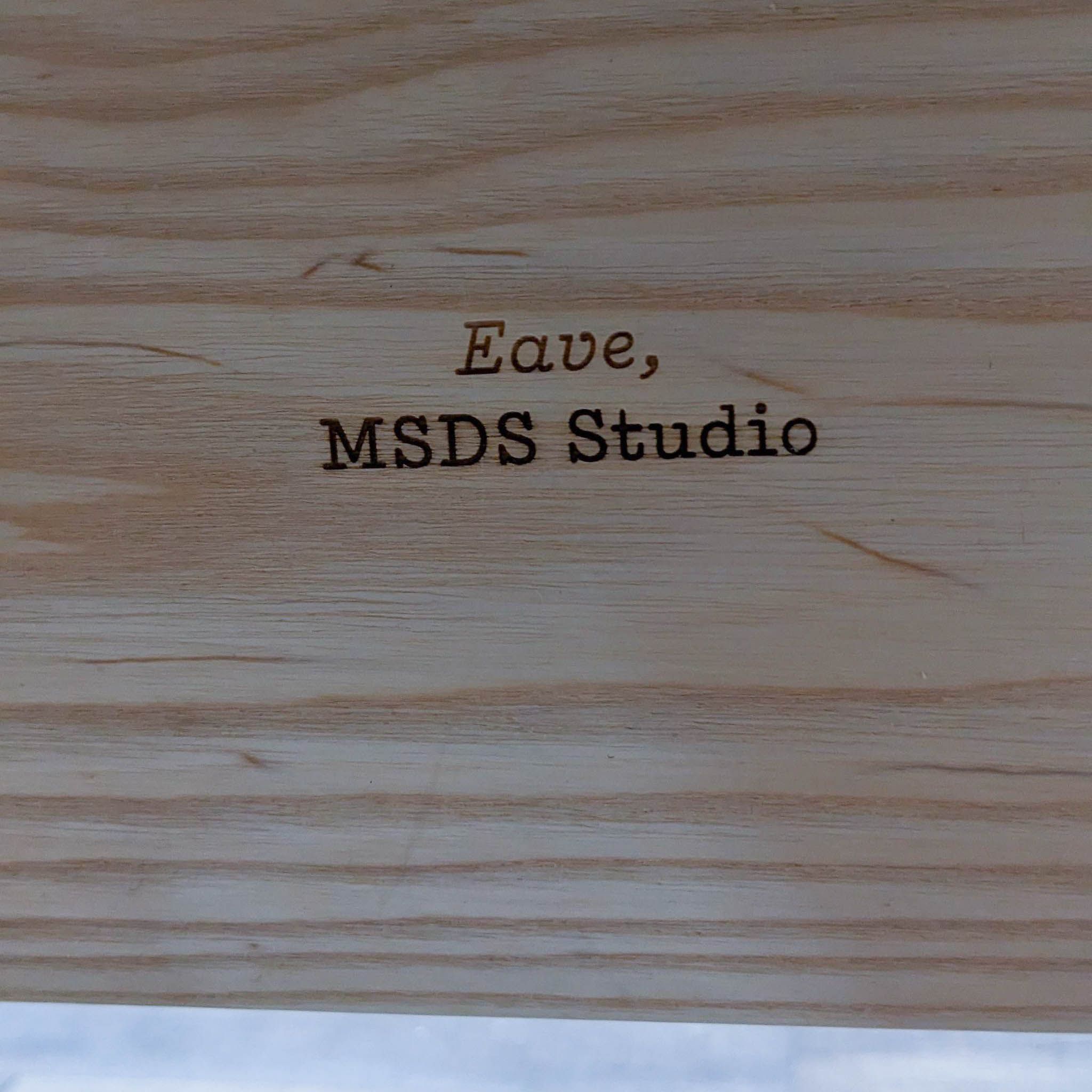Close-up of Eave desk's natural wood finish with 'Eave, MSDS Studio' branding engraved.