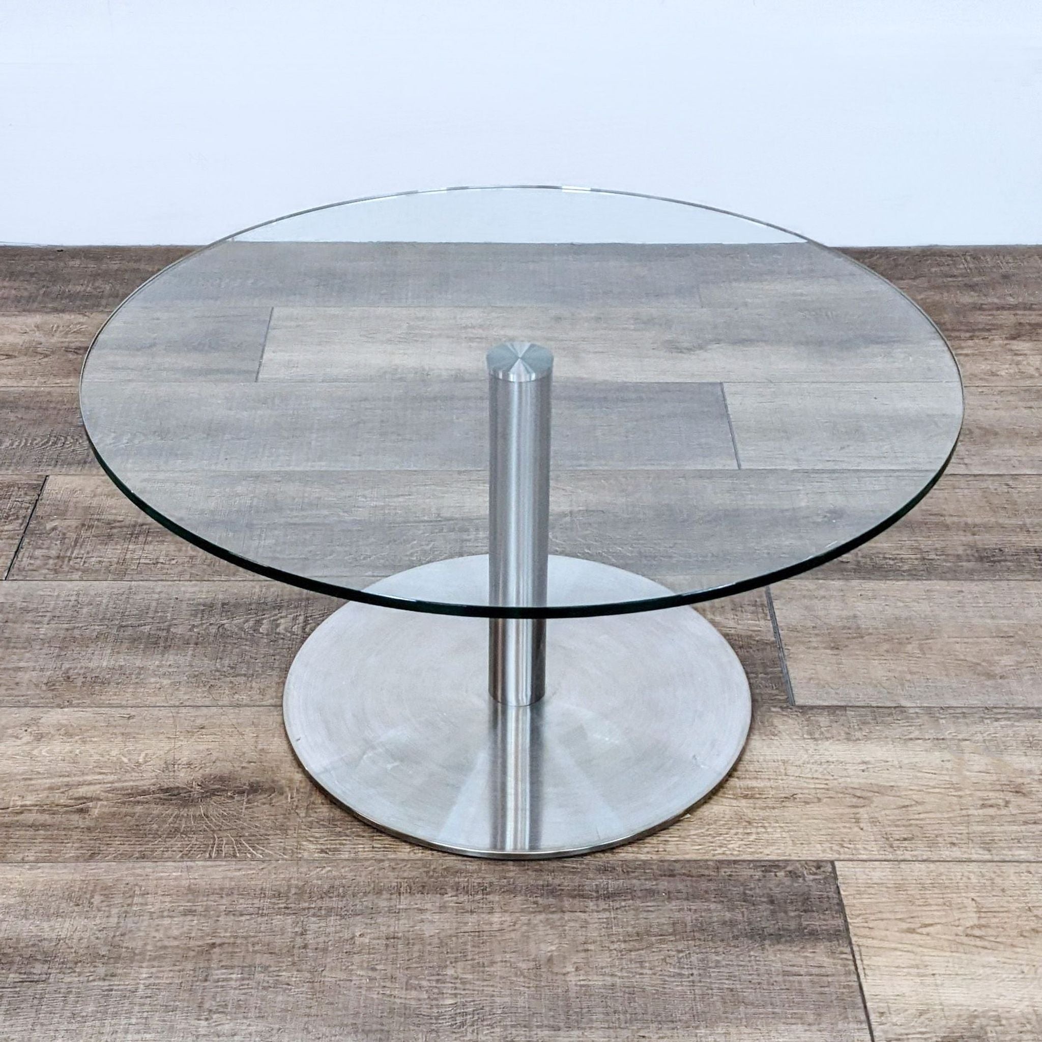 Round glass-top Reperch end table with a single metallic central leg on a wooden floor.