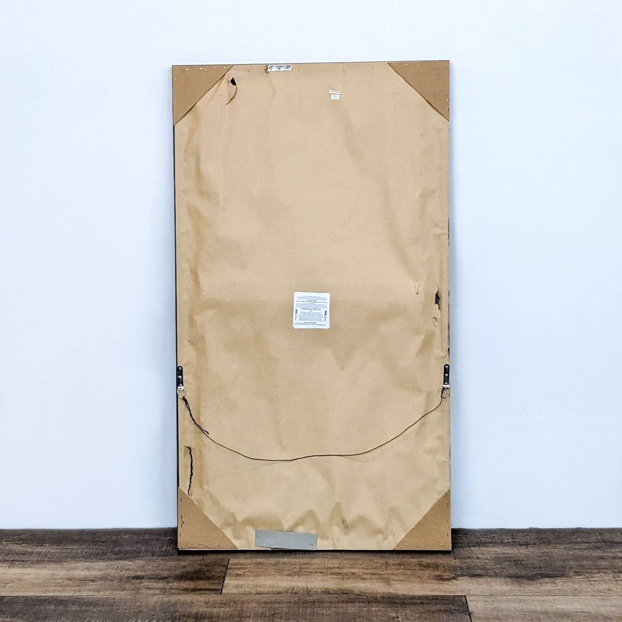 Back of a World Market rectangle mirror with paper backing and hanging hardware visible on a wooden floor against a white wall.