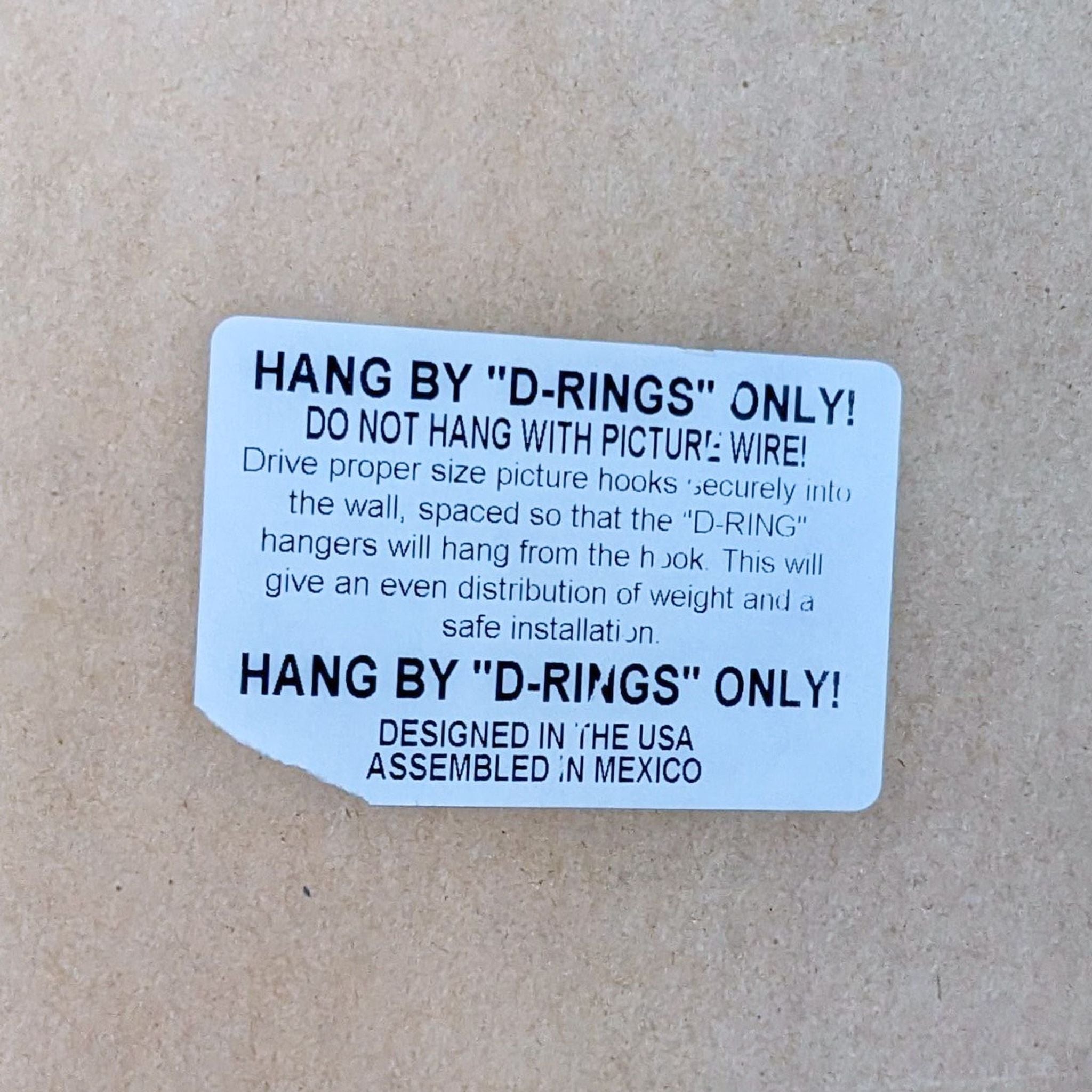Instruction label for hanging a mirror with "D-RING" only, cautioning against using picture wire, on a cardboard background.