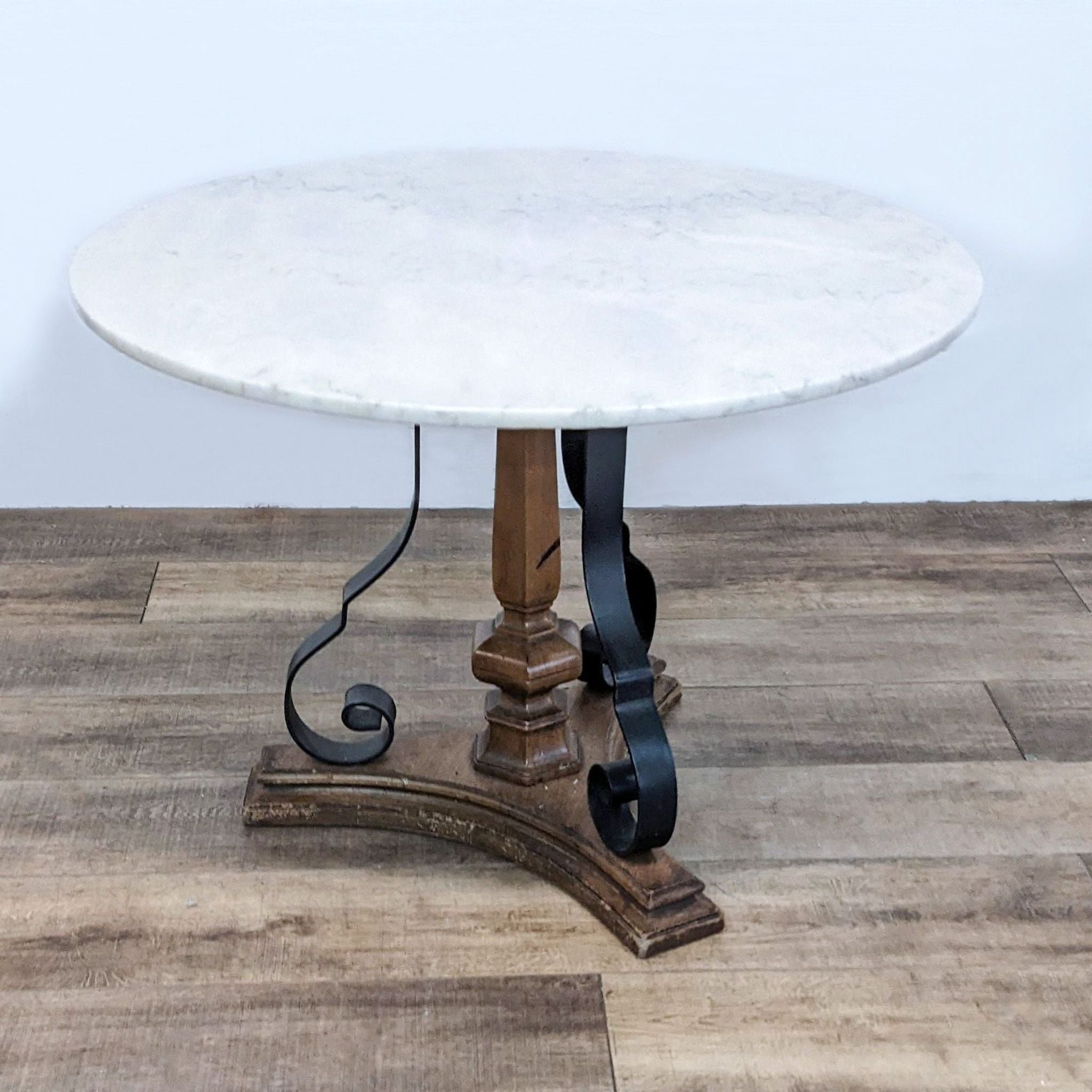 Reperch round marble-top dining table with a wooden base and metal scroll supports on wooden flooring.