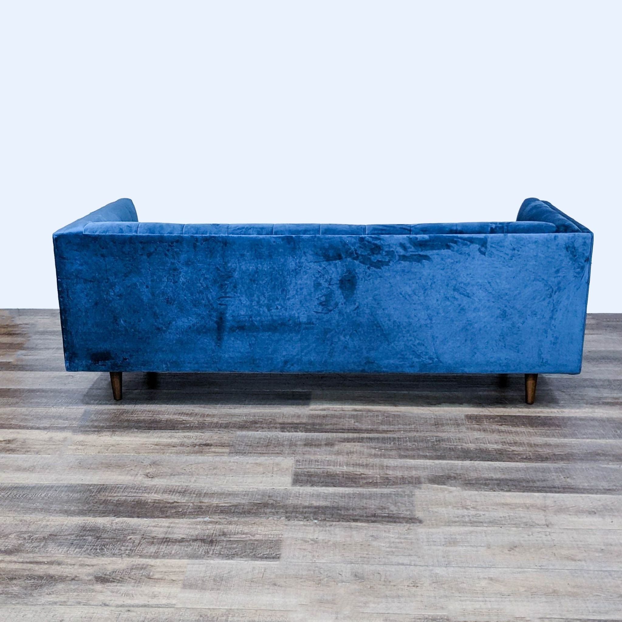 Blue velvet 3-seat West Elm sofa with tufted back and wood tapered feet, side view.