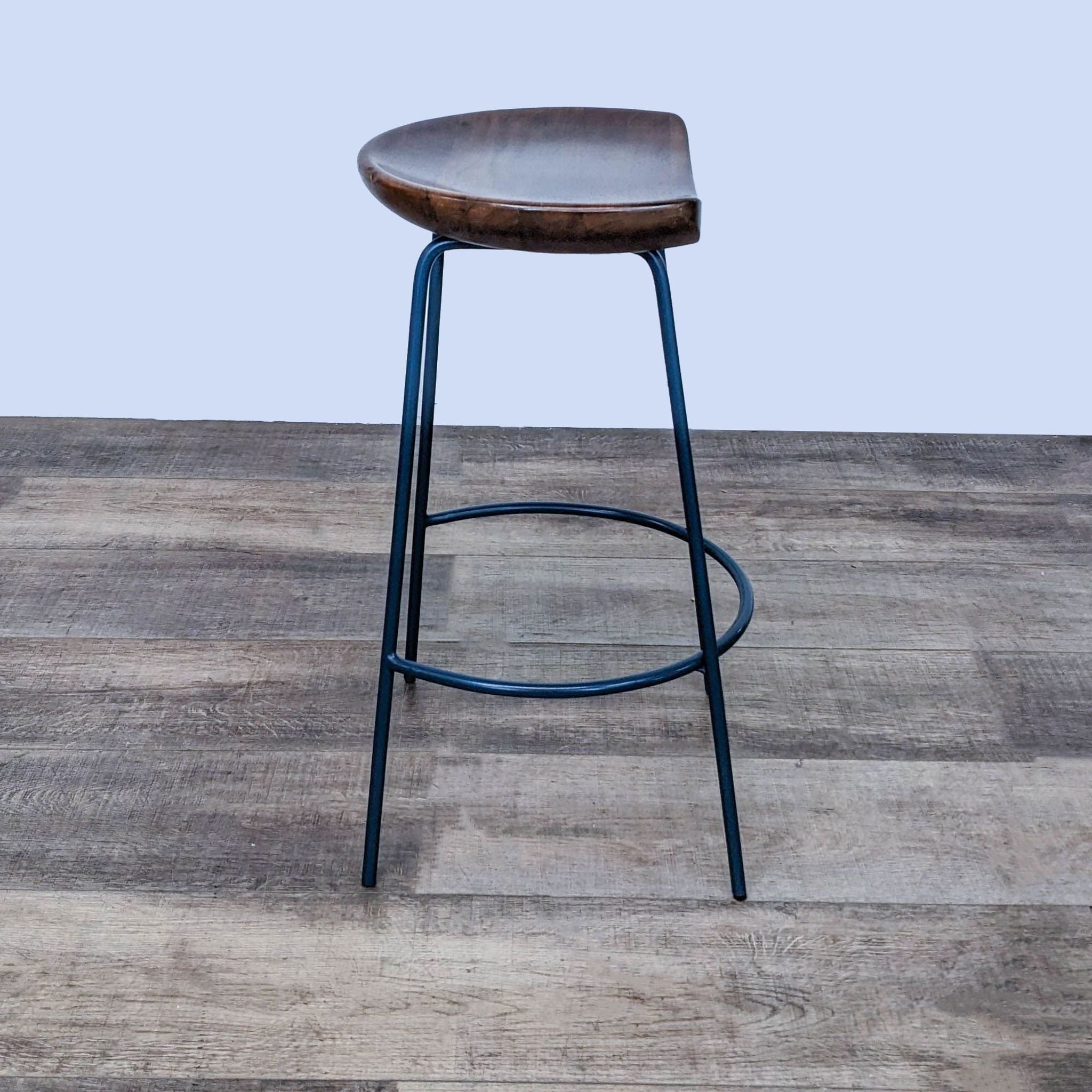 West Elm Alden Solid Wood Bucket Seat Counter Stool on a Black Steel Base, against a wooden floor backdrop.
