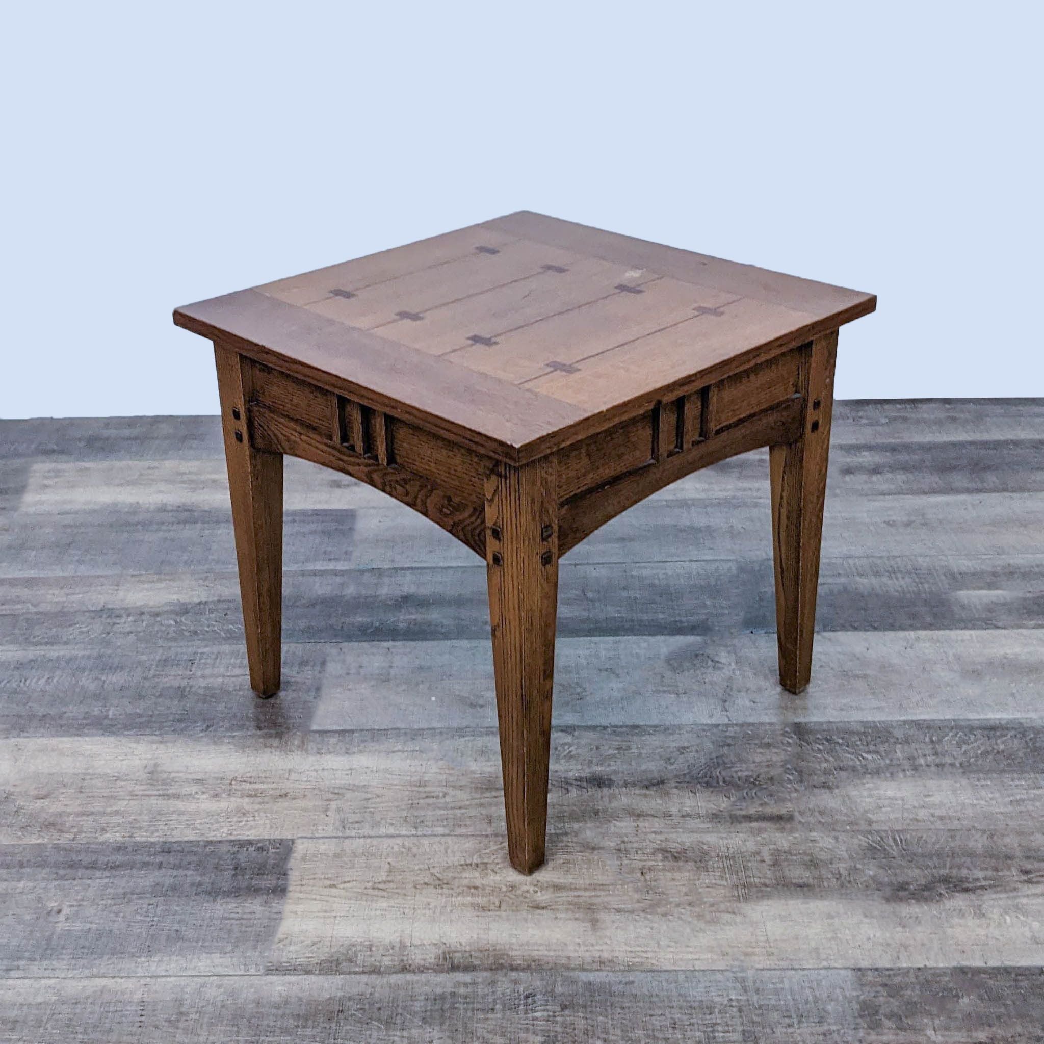 Angled view of a square oak end table by Reperch, featuring detailed inlay work, standing on a gray wood-patterned floor.