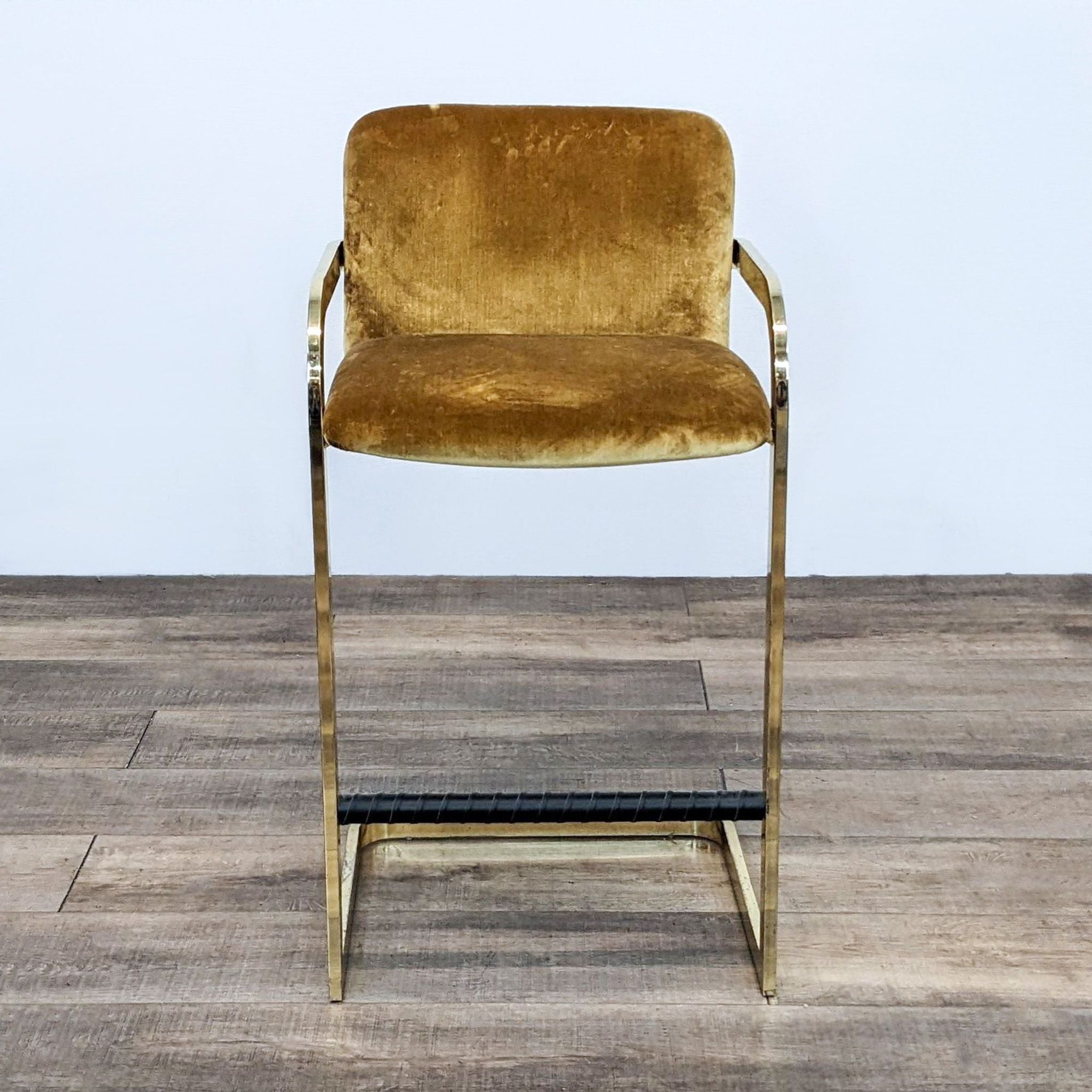 Milo Baughman gold velvet stool with brass frame on wooden floor.