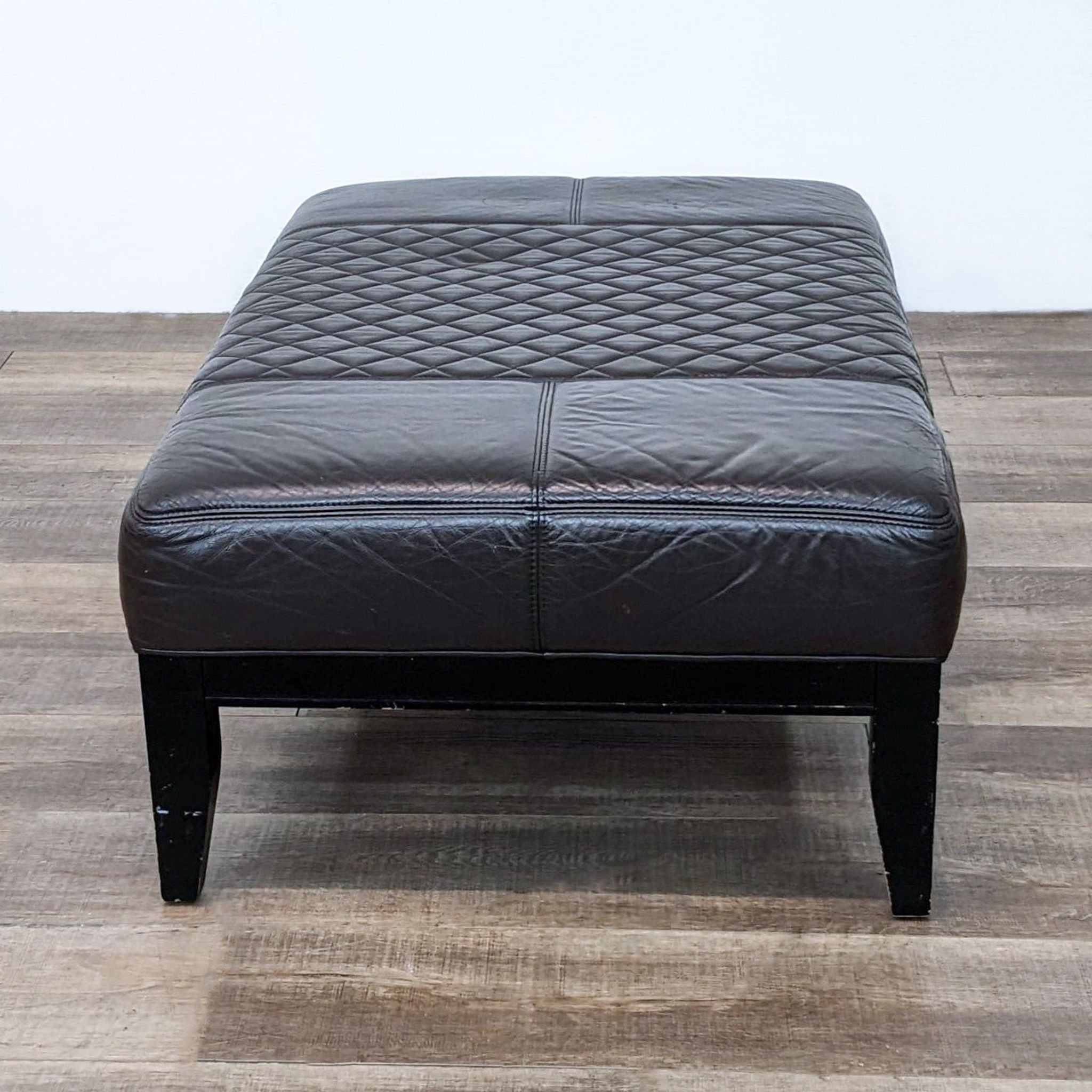 Reperch brand brown leather ottoman with quilted top and dark wooden legs on a wooden floor.