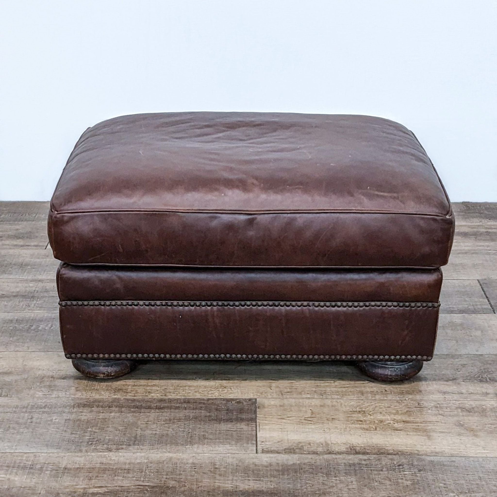Henredon Leather Co. brown leather ottoman with nail head details and wooden bun feet on a wooden floor.