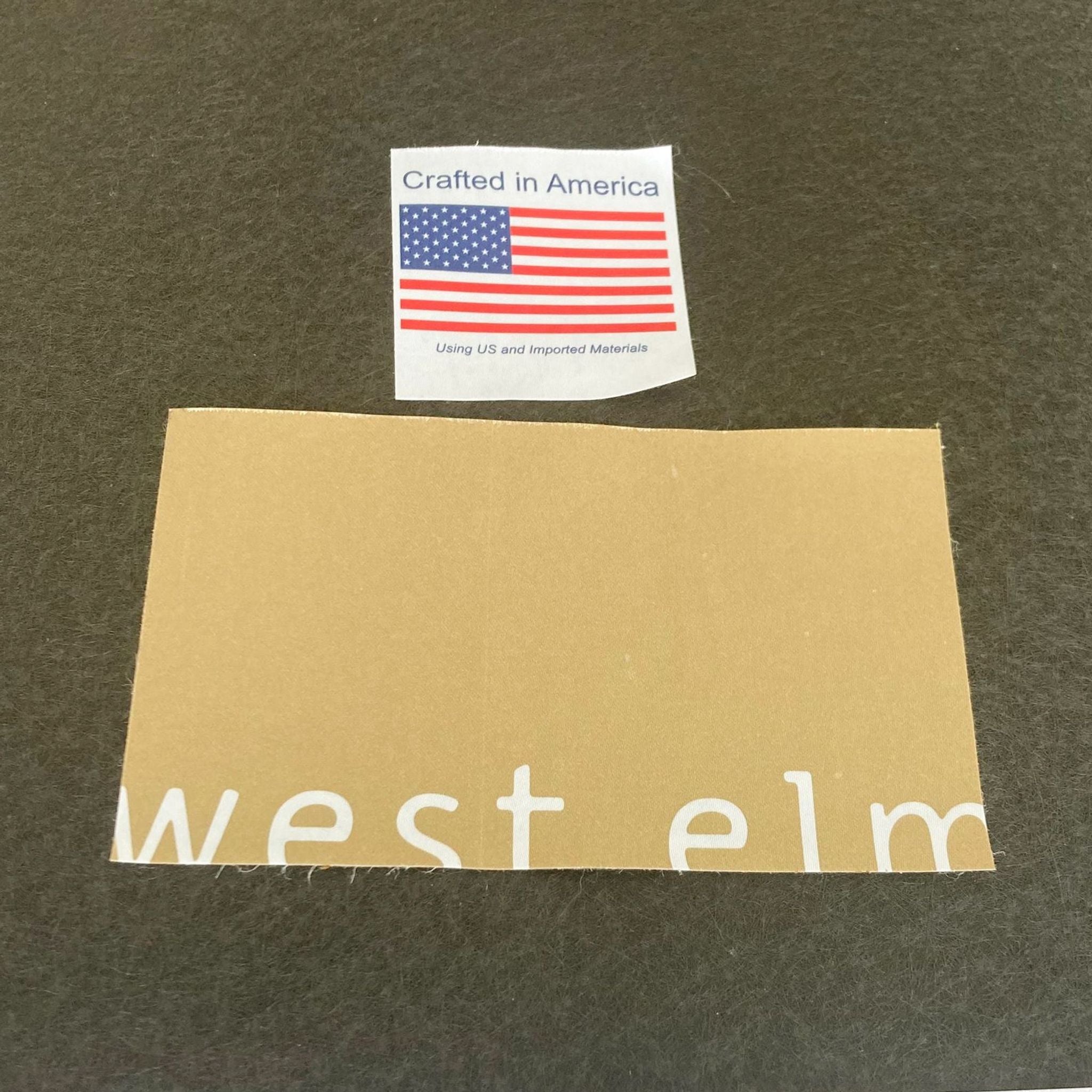 Alt text: West Elm brand label with "Crafted in America" tag on grey fabric, indicating materials used for a modern ottoman.