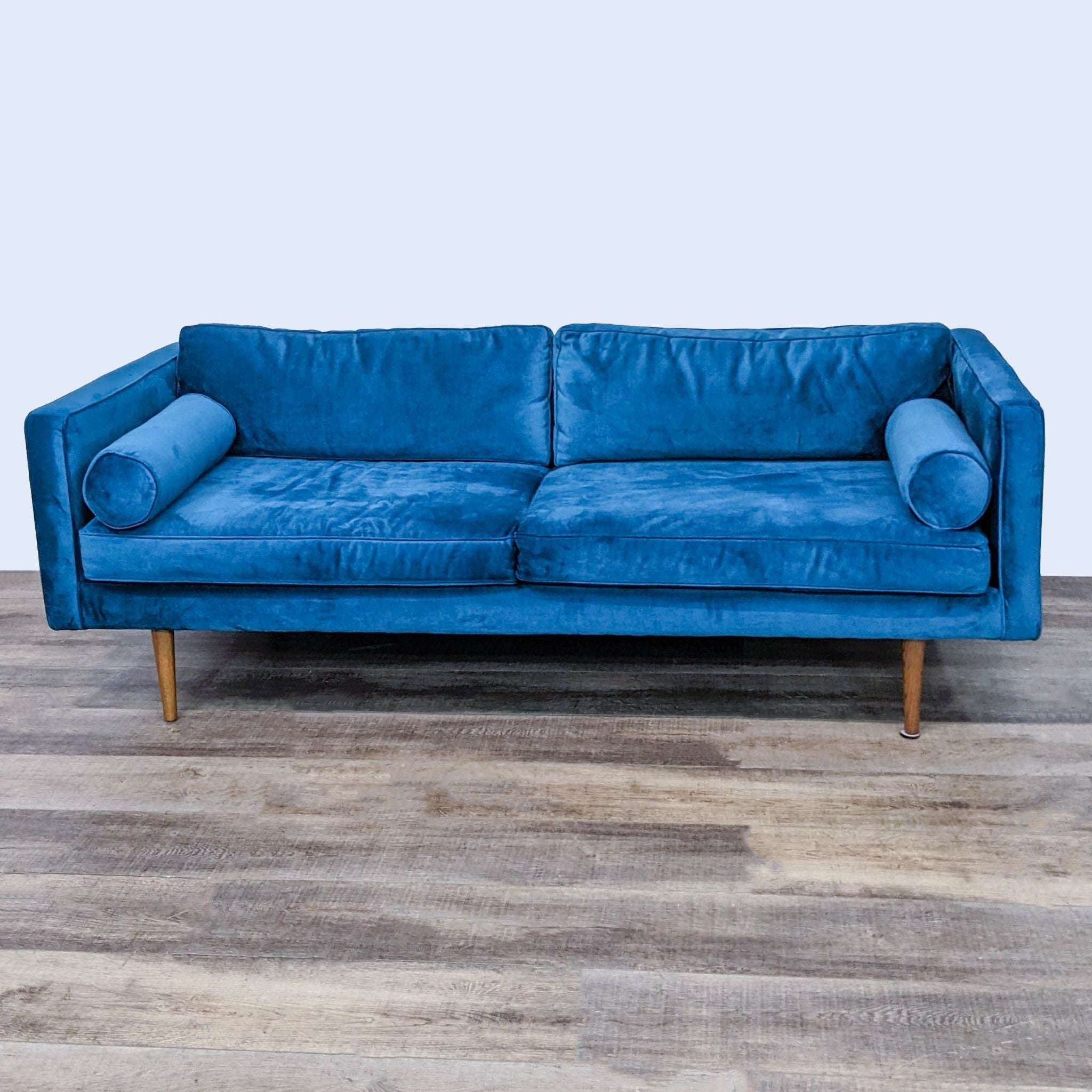 1. Blue velvet West Elm 3-seat sofa with tapered legs and two matching bolster pillows, set on a wooden floor.