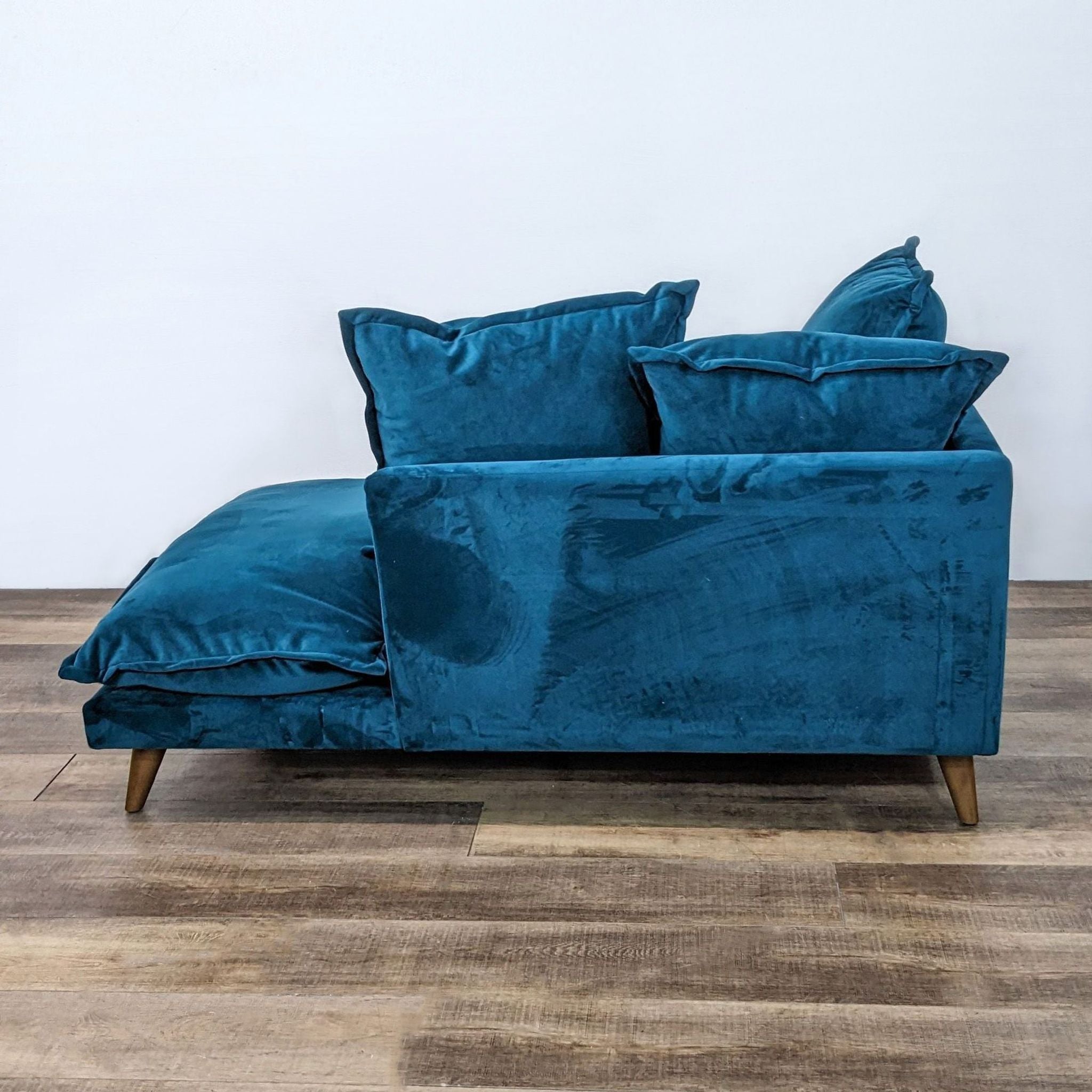Joybird Furniture's Denna chaise lounge in peacock velvet, viewed from the side, with its ottoman and wood frame with slanted legs on a wooden floor.