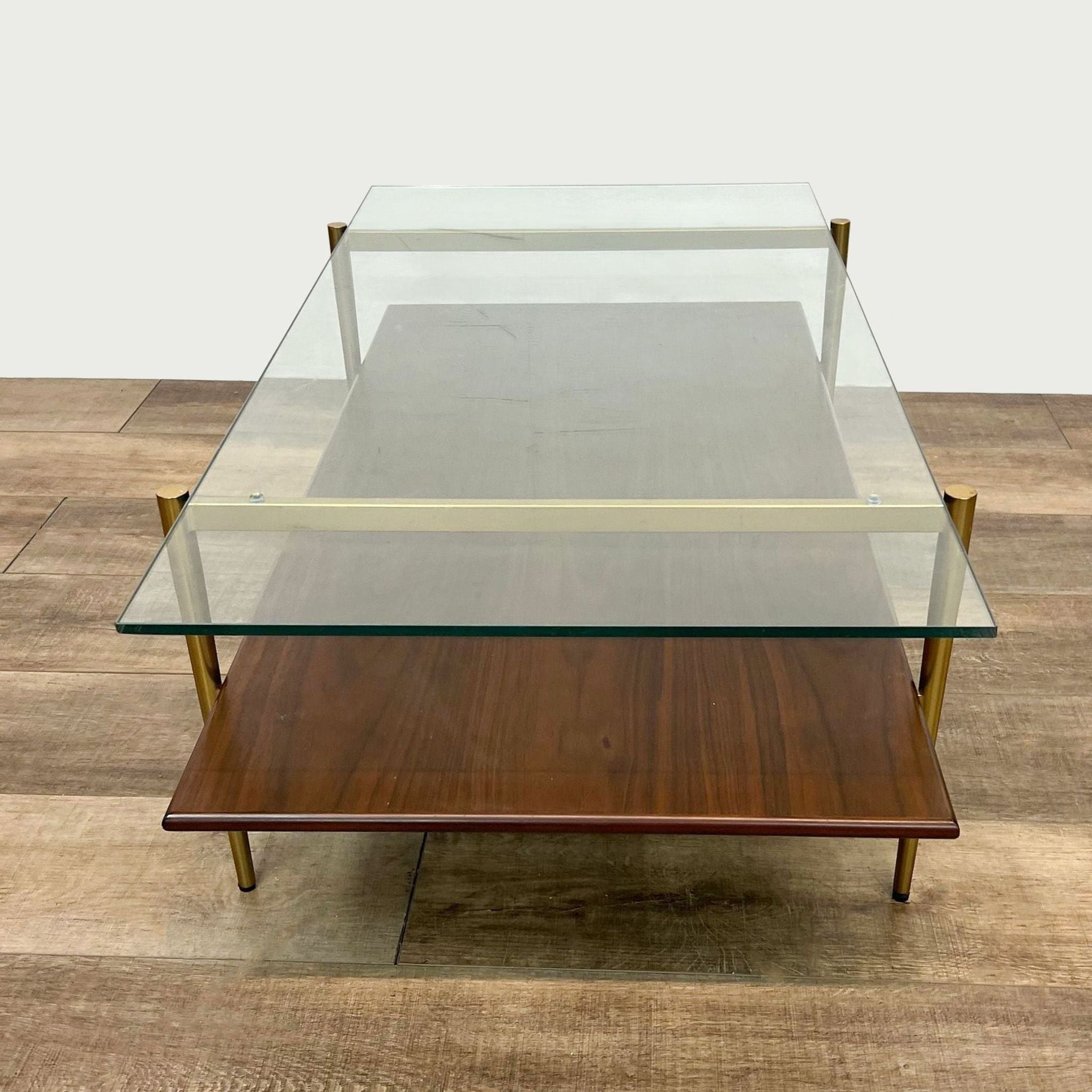 Wooden West Elm coffee table featuring a glass top layer over a lower wooden shelf with brass-finished detailing, on parquet flooring.