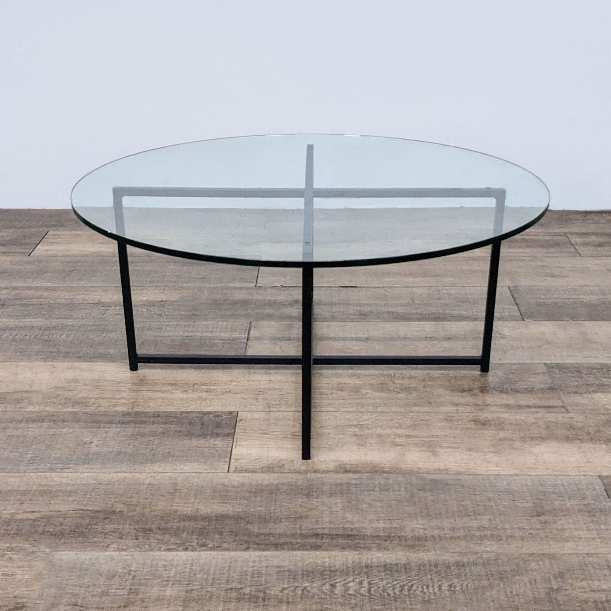 Room & Board brand coffee table with a circular glass top and minimalist black legs, set on a wooden floor.