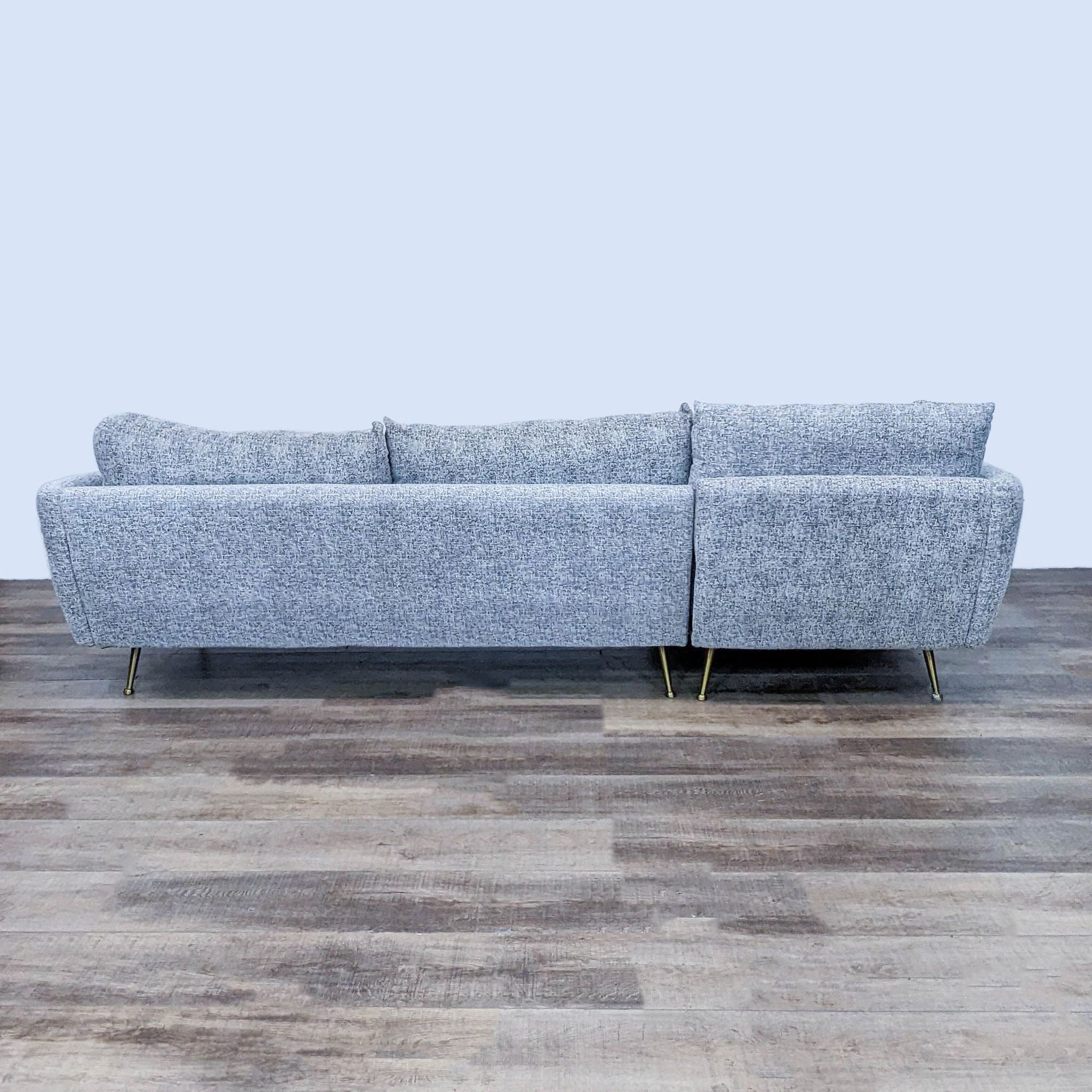 3. "Rear view of an Albany Park grey fabric sectional sofa, highlighting its simple design and sleek brass legs against a wooden floor background."