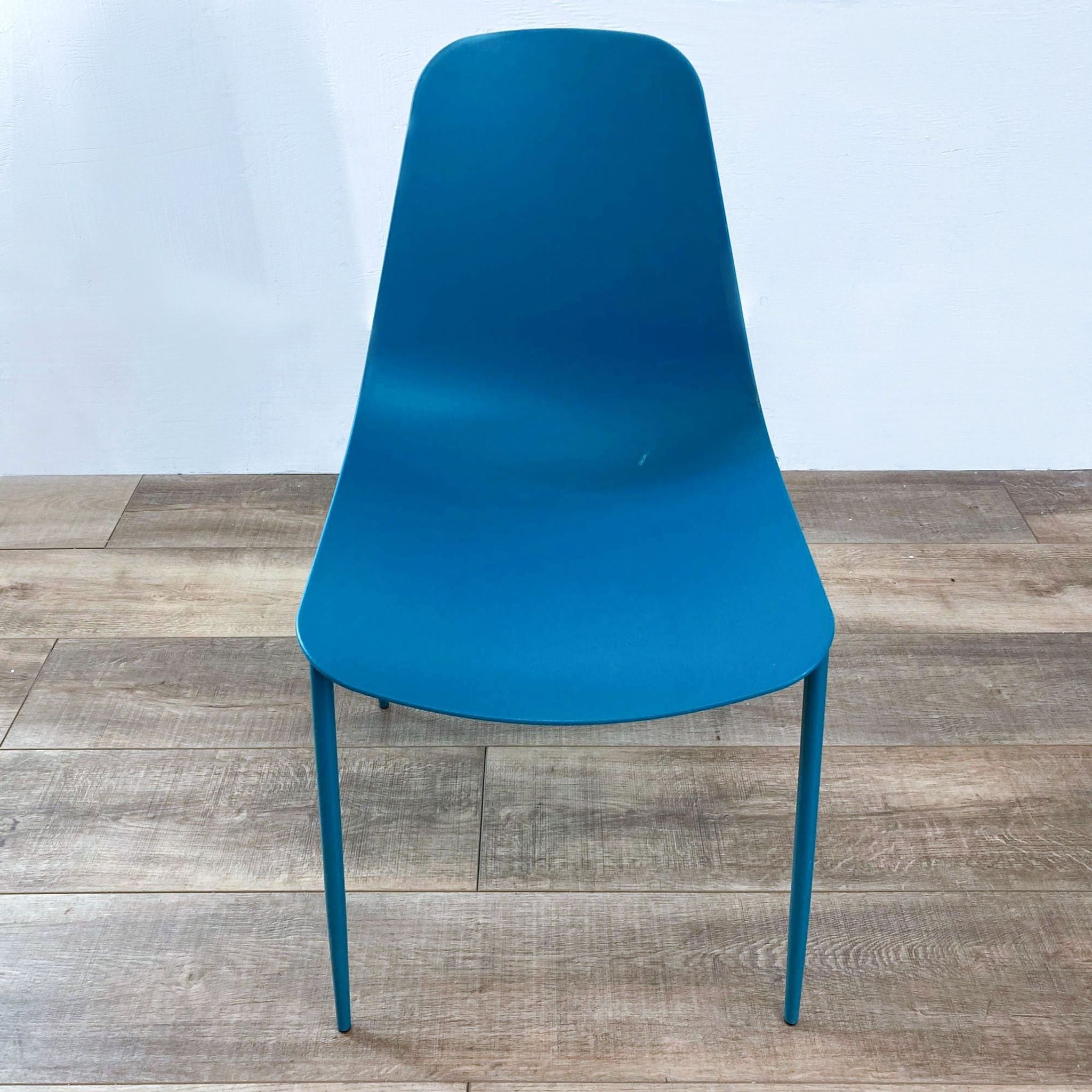 Teal plastic chair with sleek metal legs viewed from the front, against a hardwood floor and white wall.