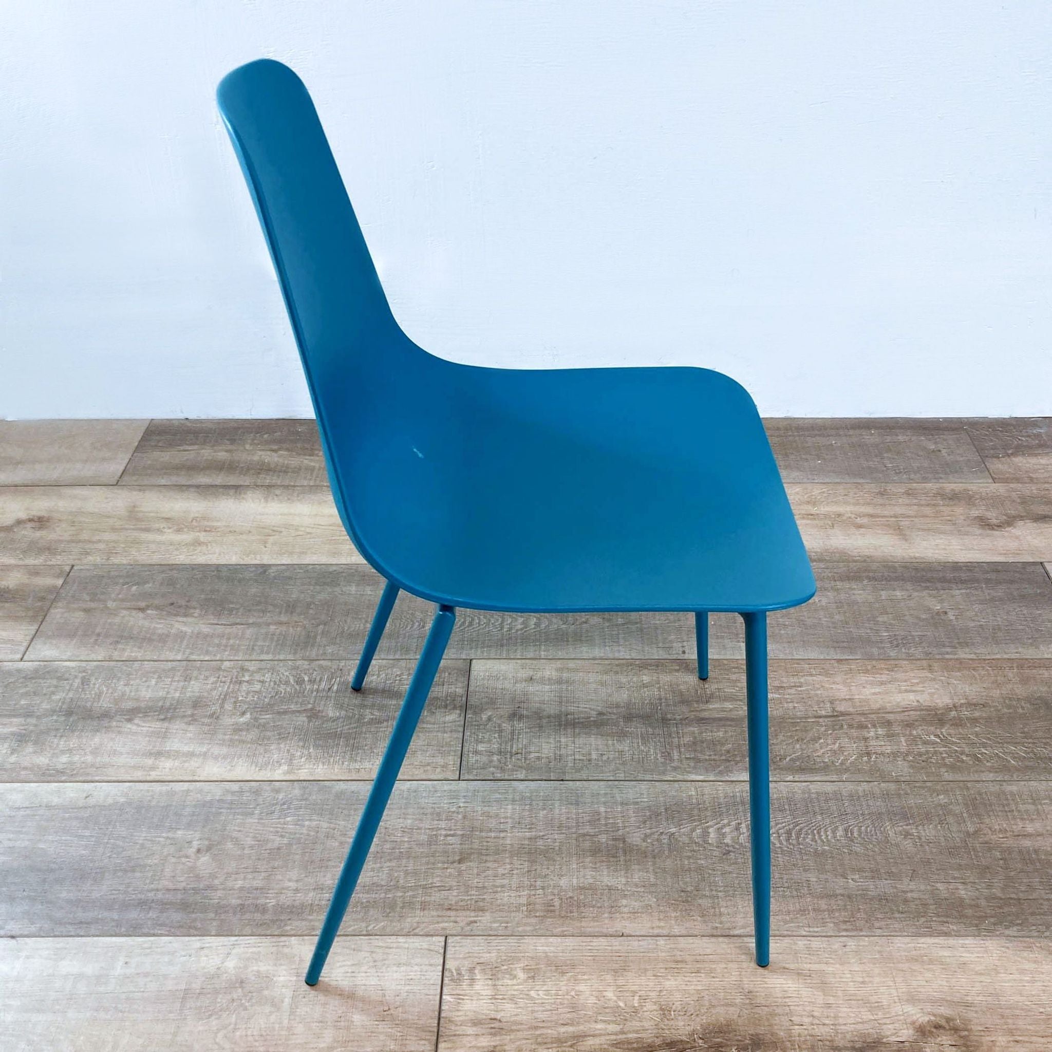 A teal Reperch modern plastic moulded chair with sleek metal legs, on a wooden floor against a white wall.