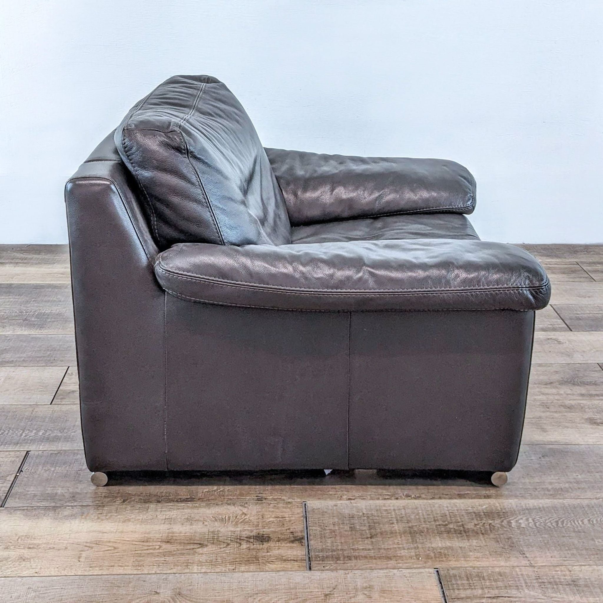 Side view of a Domicil brand leather lounge chair, showcasing its profile and wooden floor background.