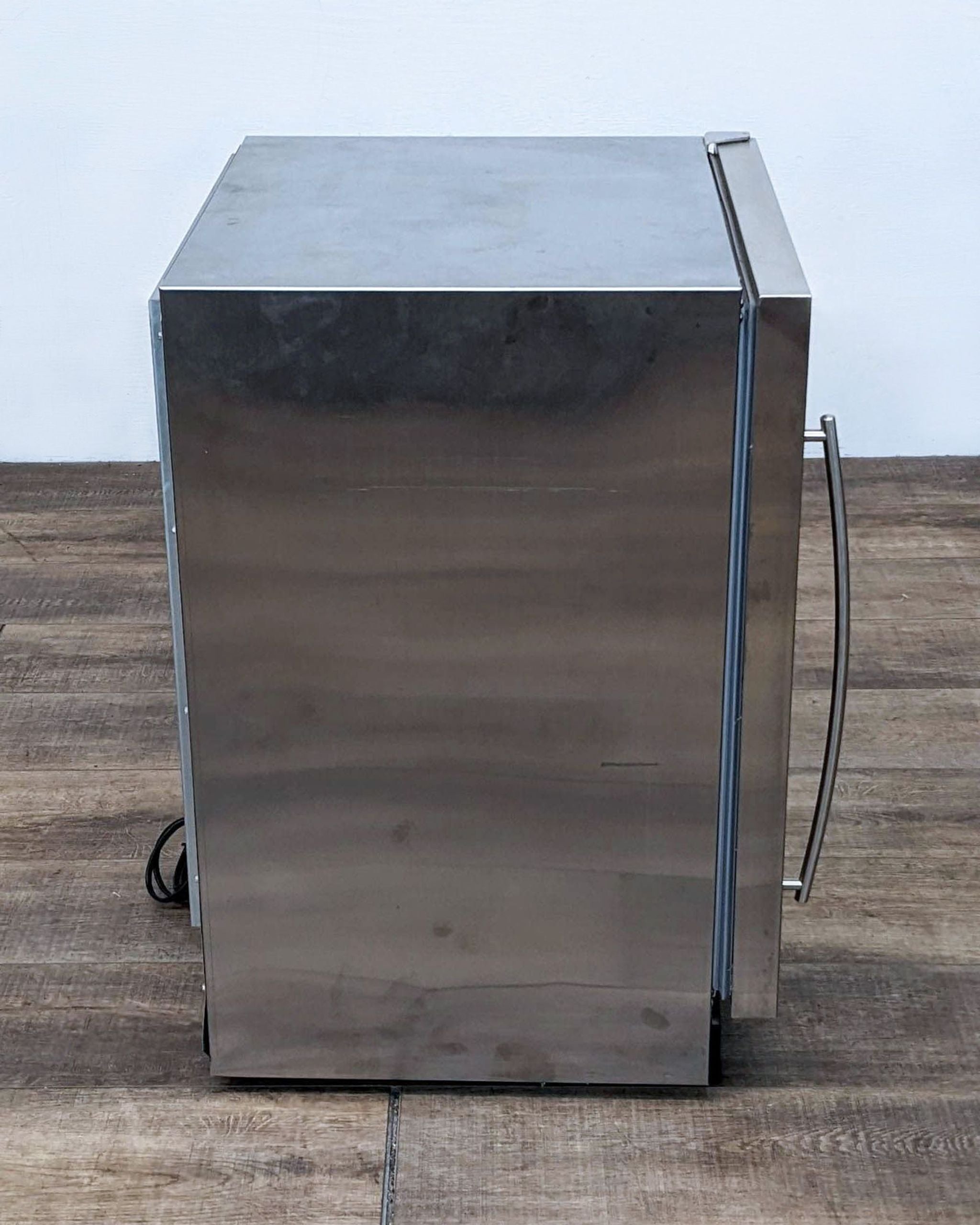 Stainless steel U-Line refrigerator with visible cord, positioned against a white wall on wooden flooring.