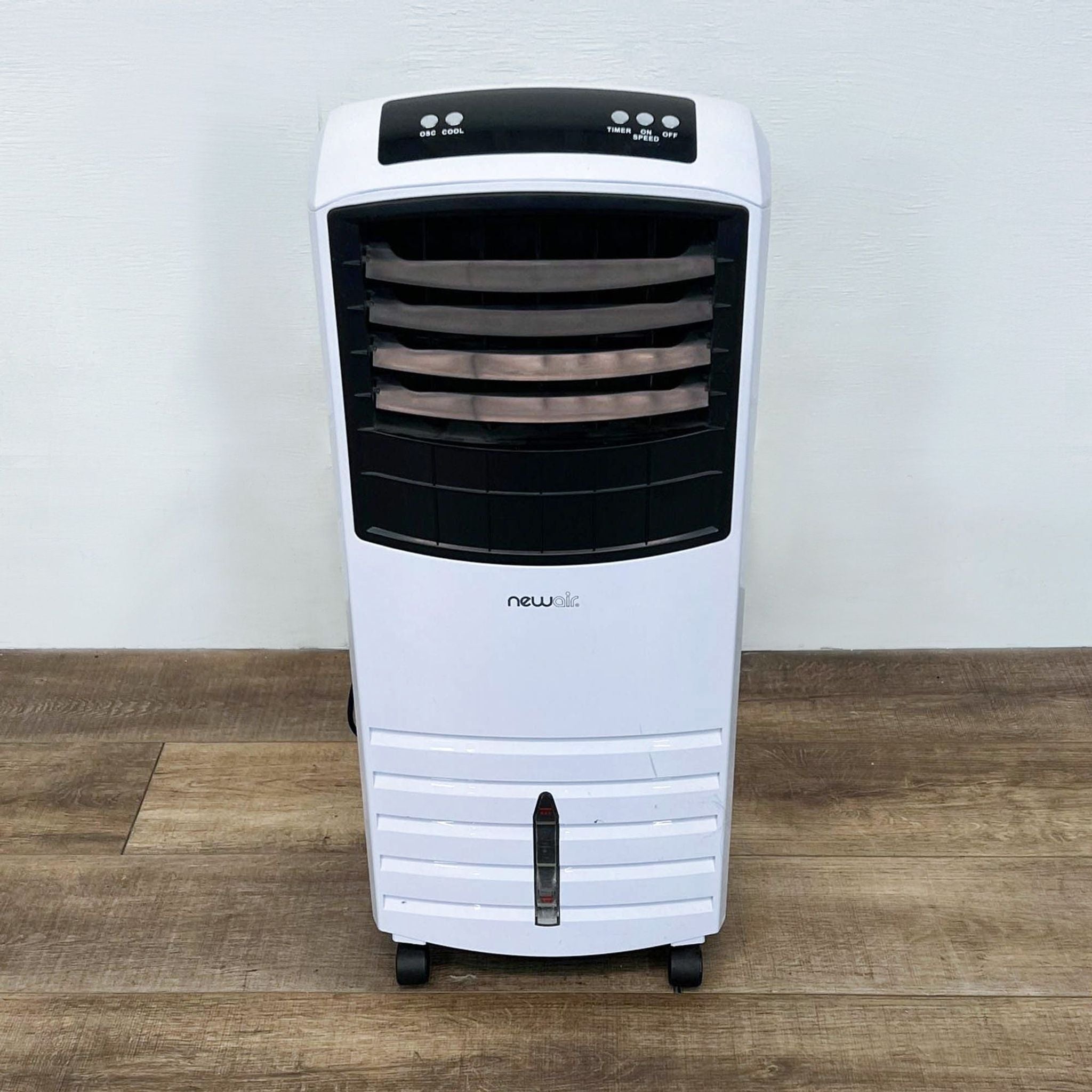 NewAir portable air cooler in white and black with control buttons visible, standing on a wooden floor against a white wall.
