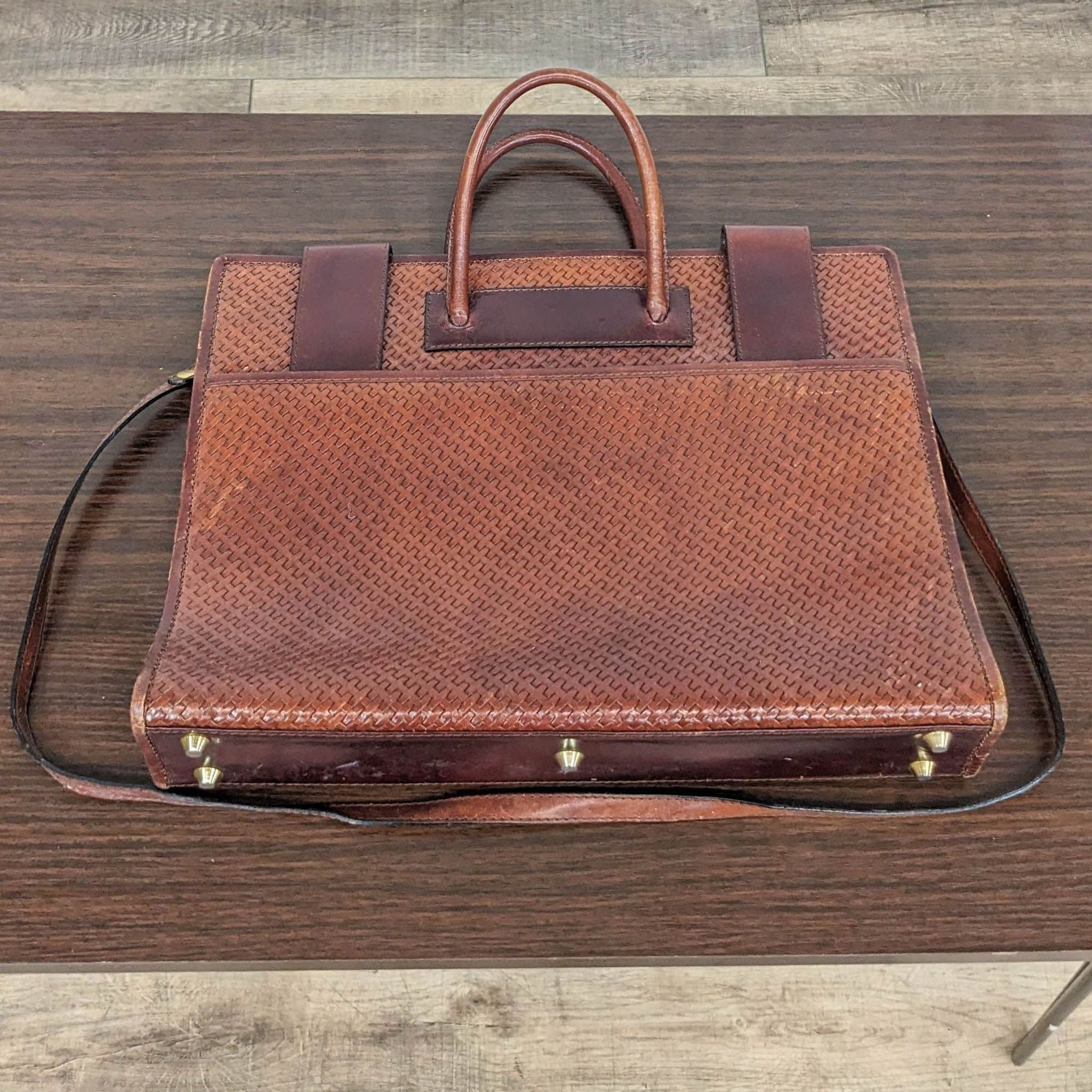 Top-down view of a textured brown Bosca leather briefcase with handles and shoulder strap on a wooden table.