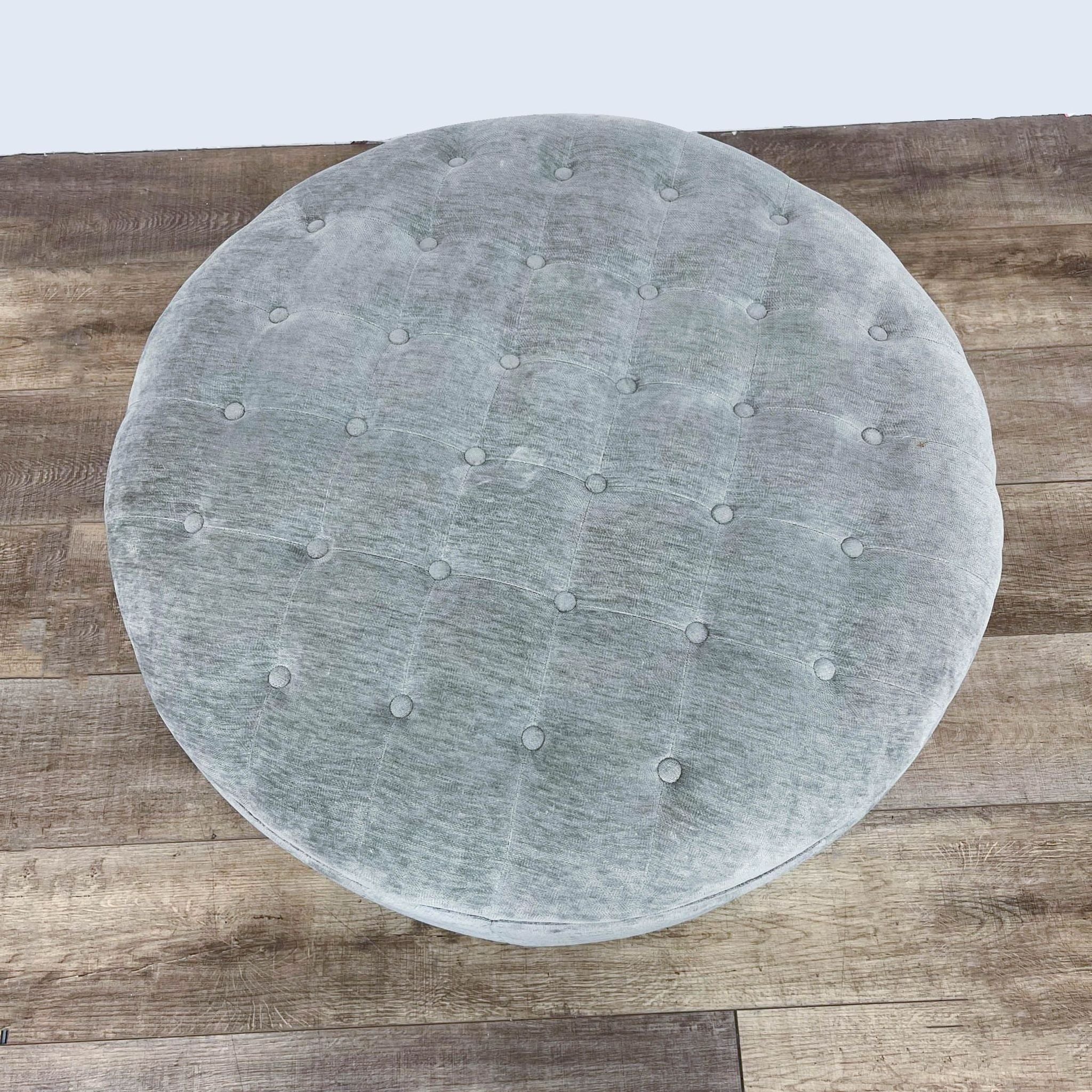 Top view of a round, tufted, dove gray 38" ottoman by Signature Design on a wooden surface.