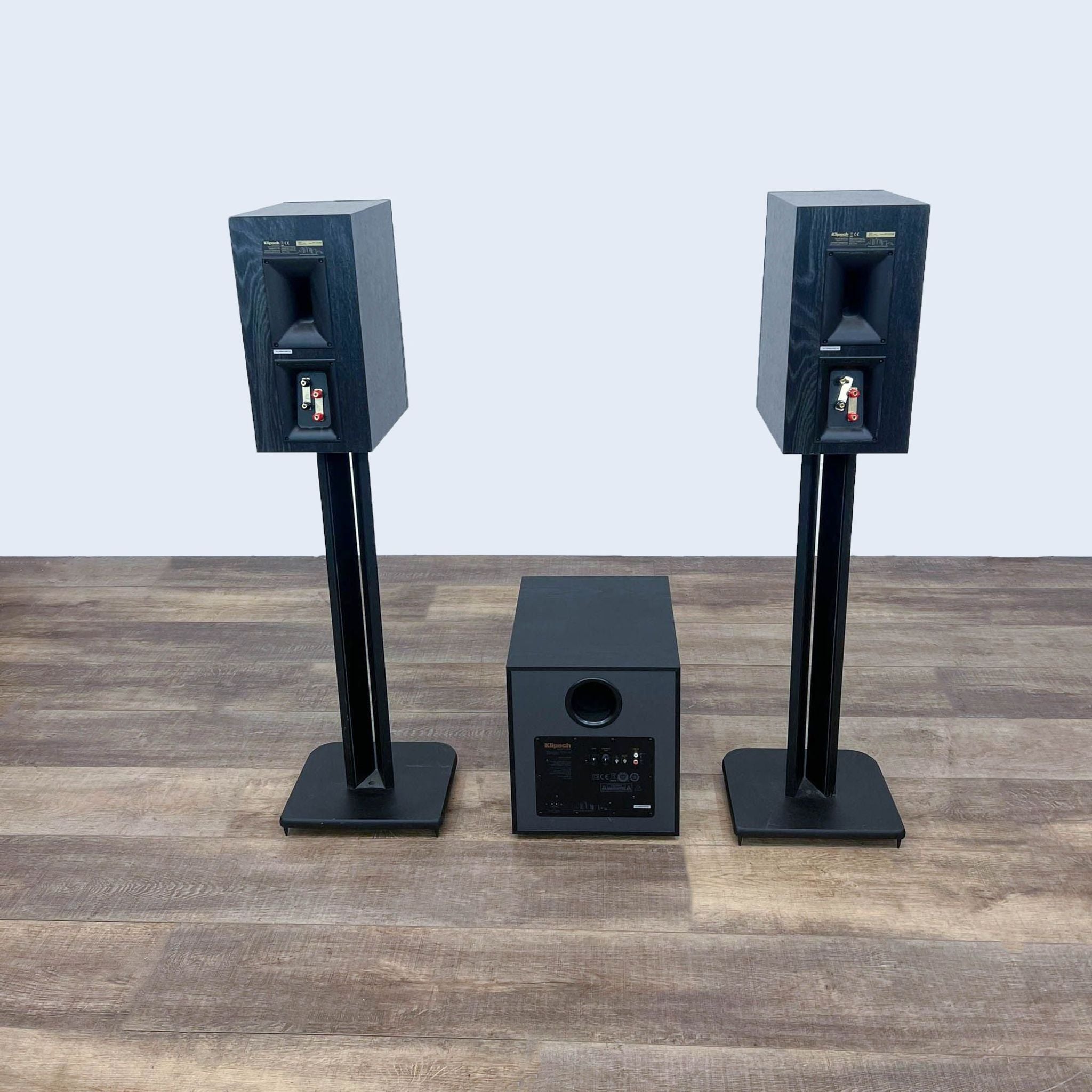 Alt text 1: Klipsch audio system with two tall black speakers on stands and a central subwoofer on a wood floor against a gray background.