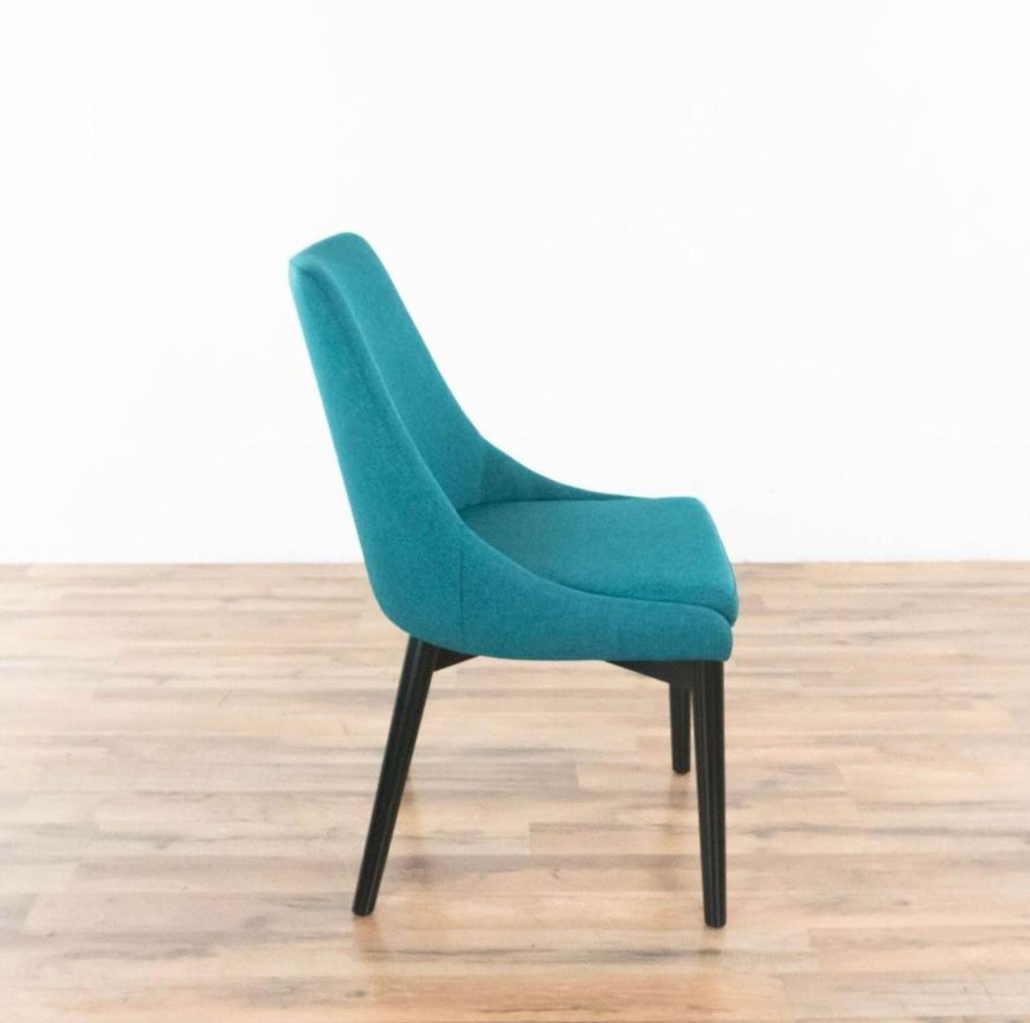 Side angle of a contemporary teal upholstered StealMod dining chair with black wooden legs, showcasing its profile.