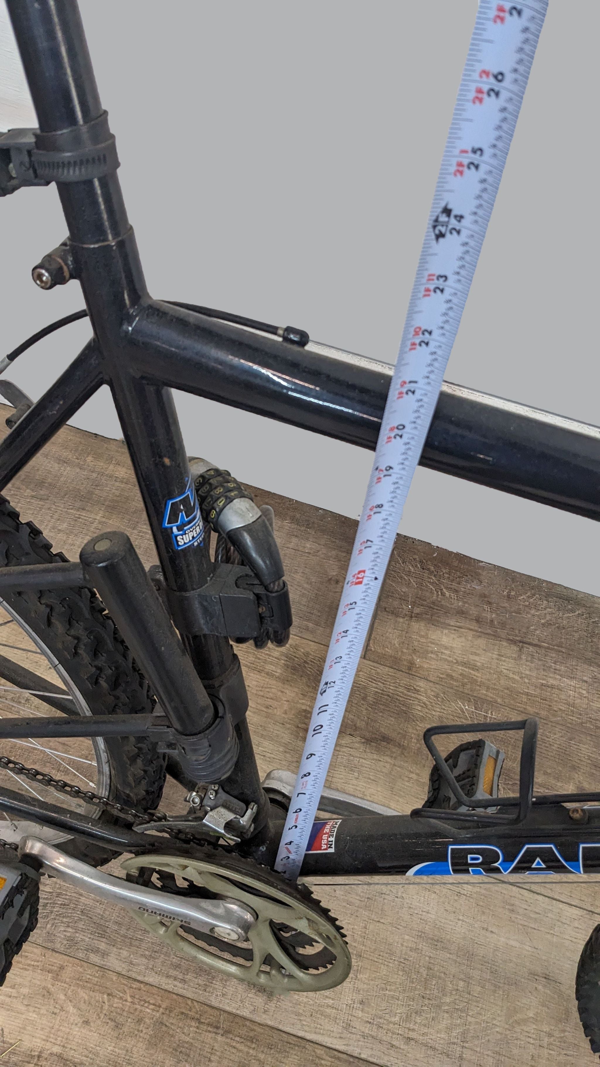 Black Raleigh mountain bike with a visible measuring tape against the frame, displayed on a wooden floor.